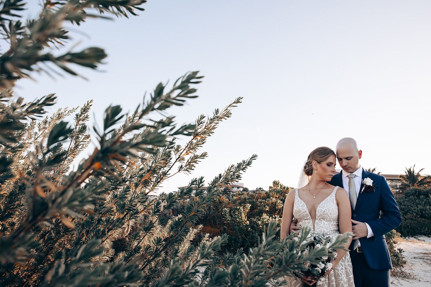 A bride and groom stand in a field of tall grass and flowers at sunset.