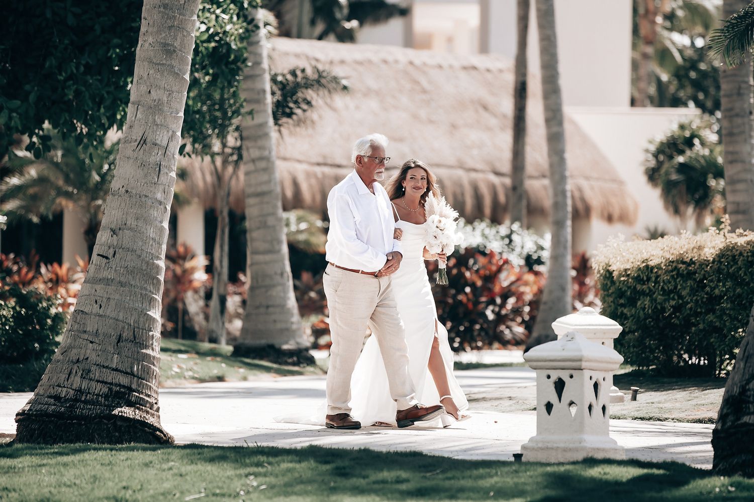 A couple in formal attire stand together on a paved path surrounded by palm trees.