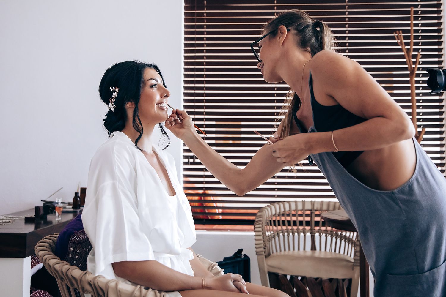 A woman applying makeup to another woman's face in a cozy indoor setting.