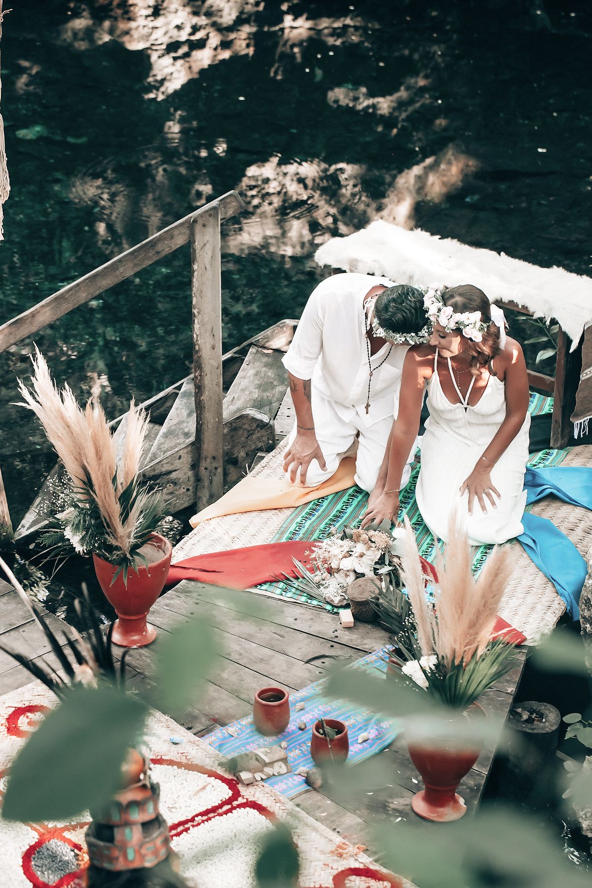 A couple stands on a wooden platform overlooking a lush, forested landscape with a body of water.