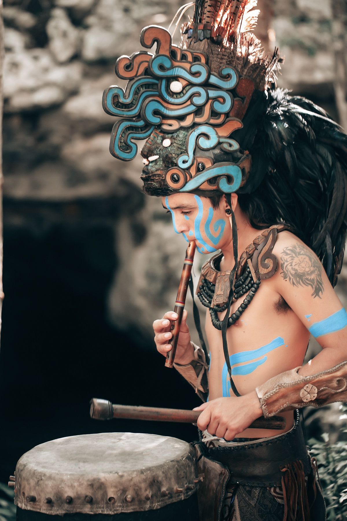 An indigenous person wearing an elaborate headdress and holding a drum in a natural setting.