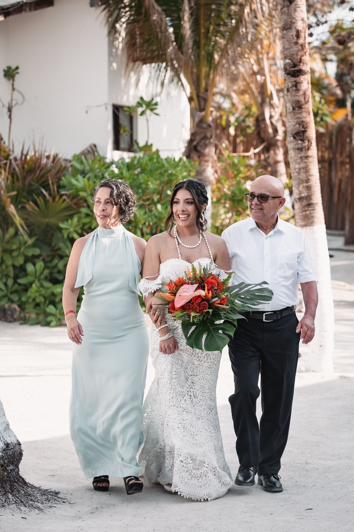 Bride in lace wedding dress with tropical bouquet walks with parents on sandy beach.