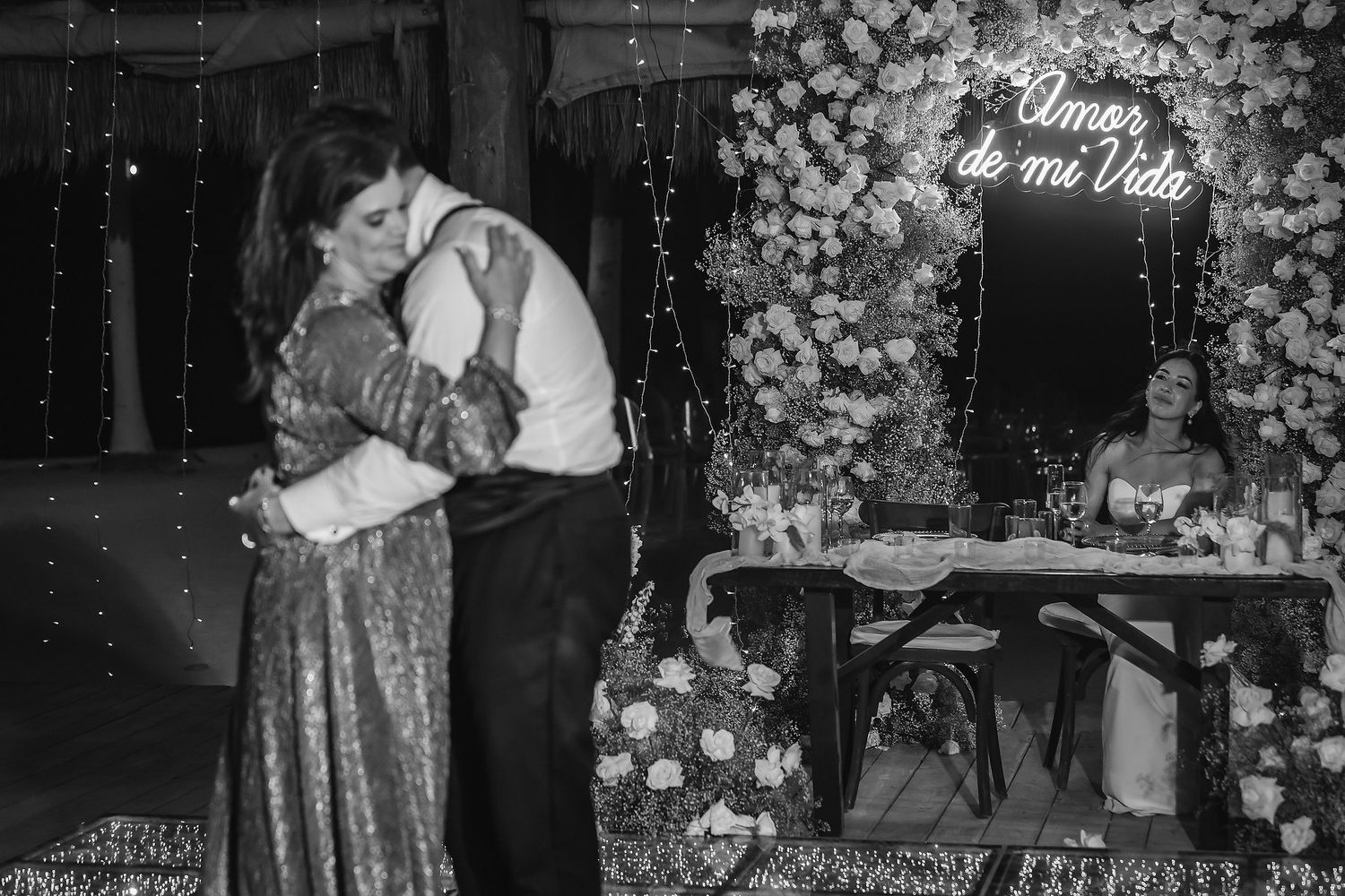 Black and white wedding photo of couple embracing on dance floor with floral backdrop and neon sign reading Amor de mi Vida.