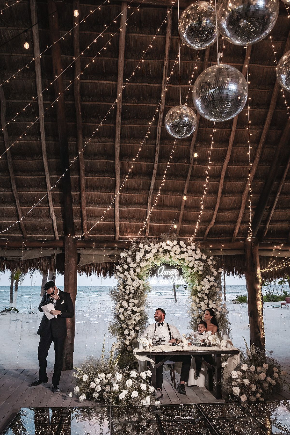 Beach wedding reception under thatched roof with disco balls, fairy lights, floral arch, and couple at sweetheart table.