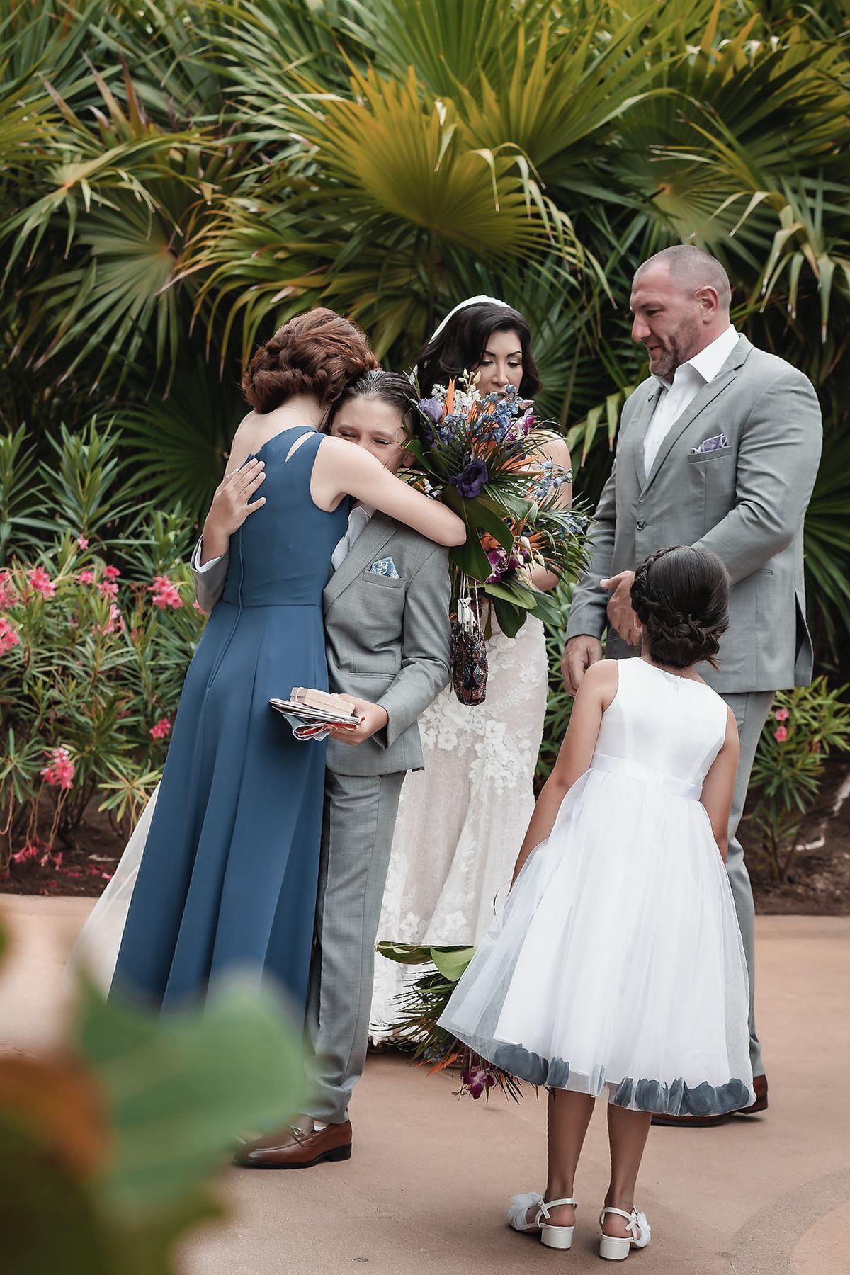 Wedding party embracing after ceremony outdoors, bride holding bouquet, flower girl in white dress, tropical garden backdrop.