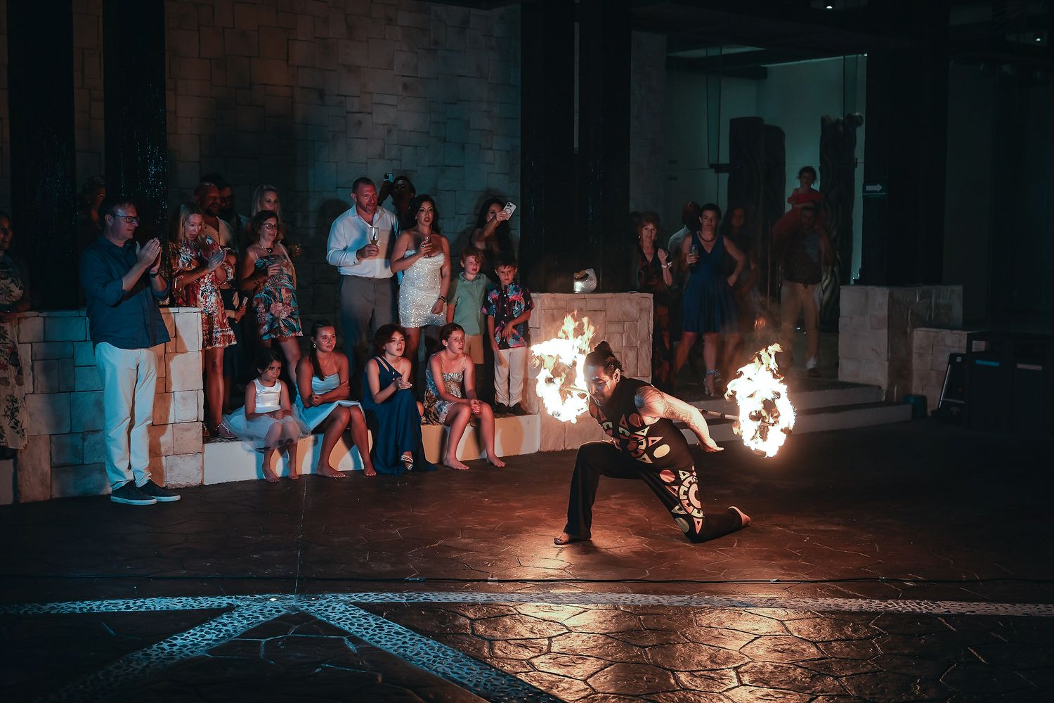 Fire performer spinning flames at a live outdoor event with an audience watching in the background at night.