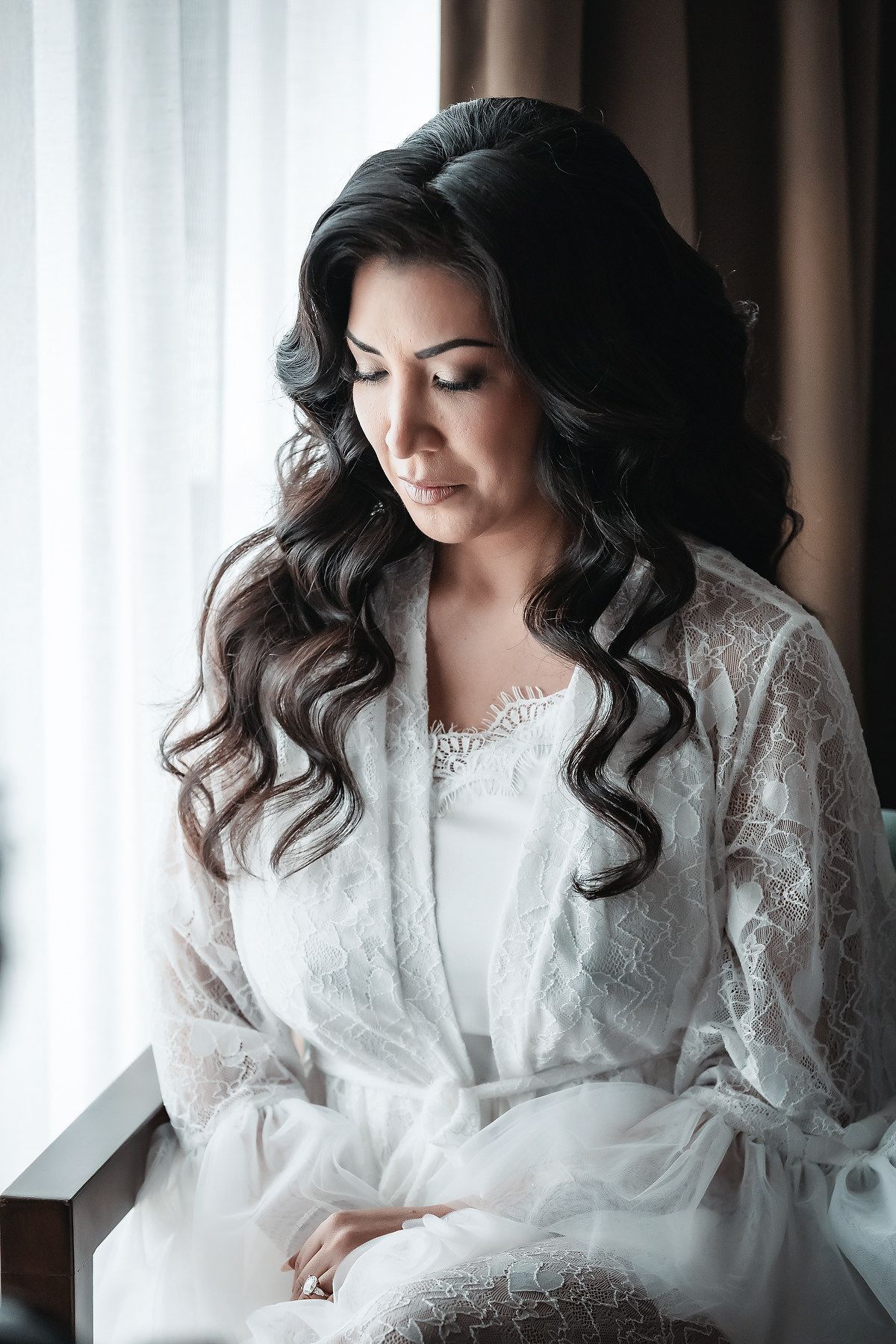 A bride with dark curly hair sits gracefully in a white lace robe near a window, gazing downward.