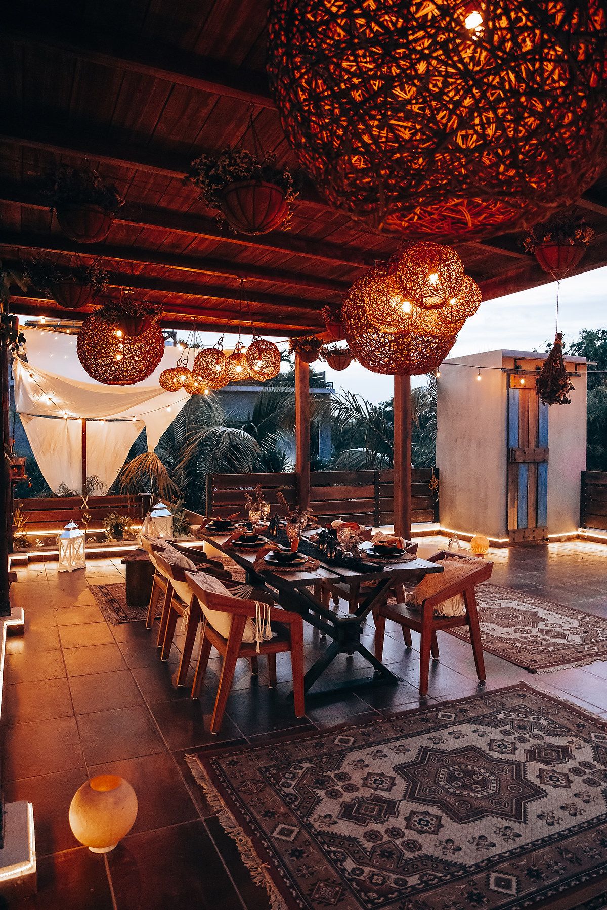 Warmly lit outdoor dining area with large wicker pendant lights, wooden table, and Persian rug at twilight.