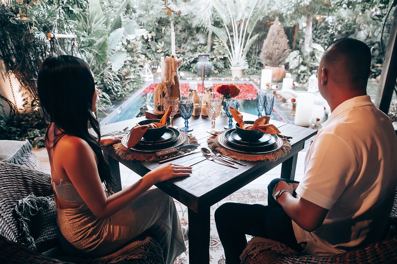 Couple enjoying a romantic outdoor dinner at a beautifully decorated table with black plates in a tropical garden setting.