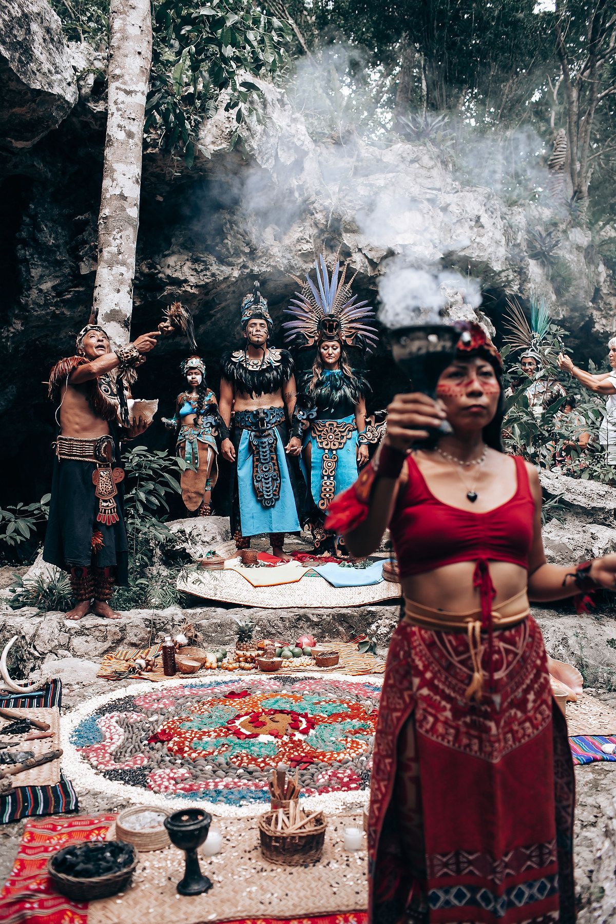 Mayan ceremony with costumed performers and ritual offerings arranged on colorful textiles amid rising incense smoke.