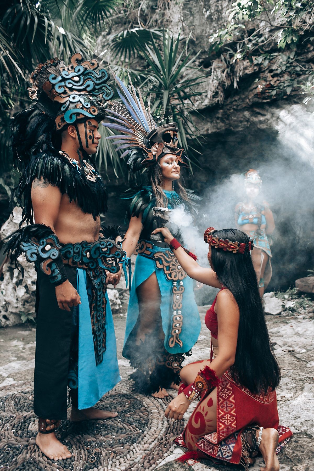 Mayan performers in traditional turquoise and feathered costumes conduct a sacred ritual ceremony in a jungle cave setting.