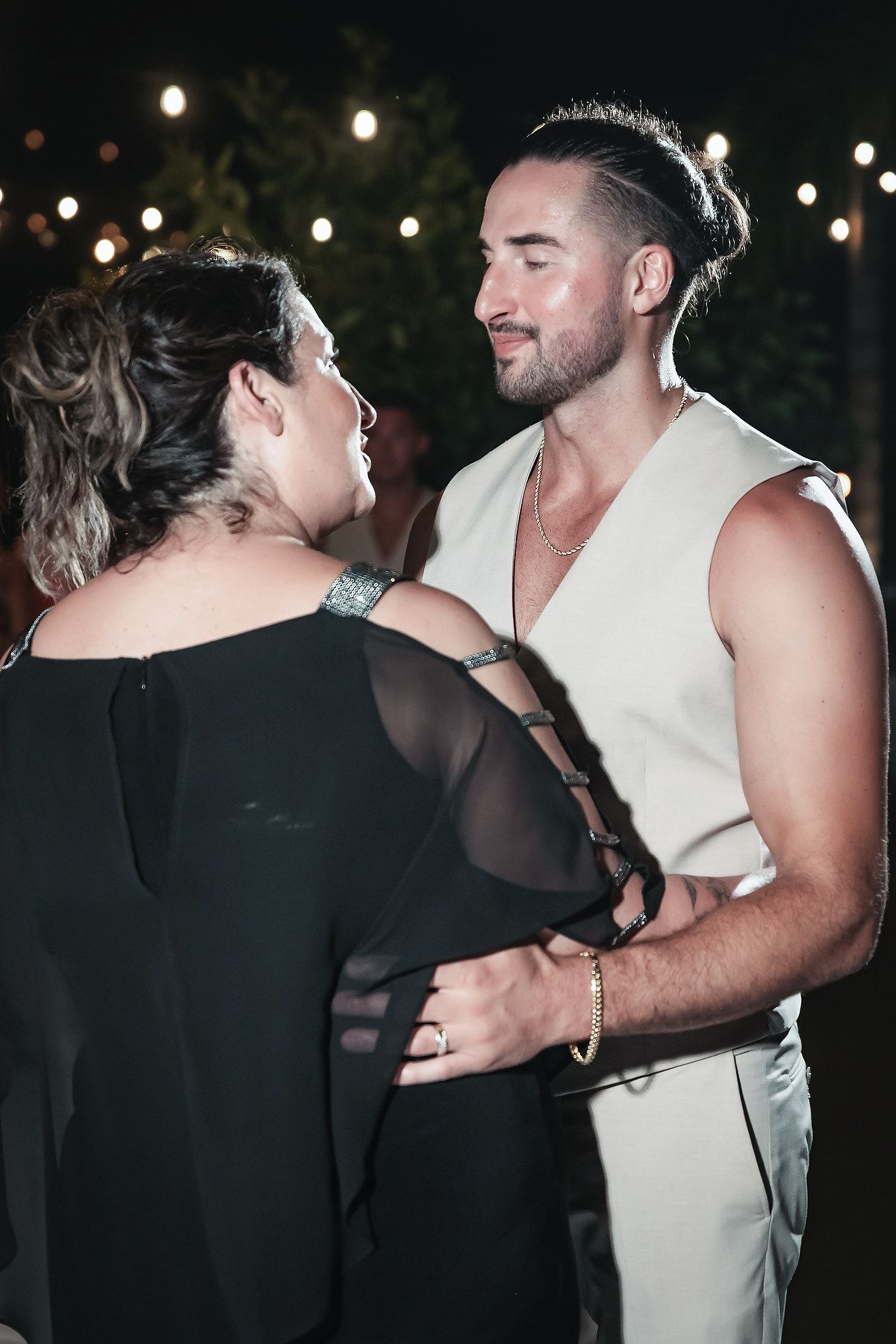 Couple dancing closely at an outdoor evening event with string lights, man in white vest and woman in black dress.