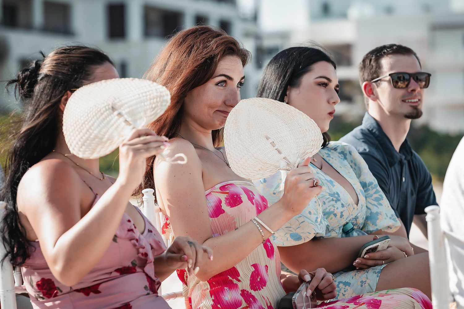Wedding guests in floral dresses fanning themselves with white paper fans at an outdoor ceremony.