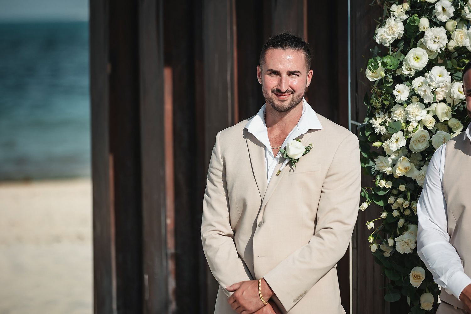Smiling groom in beige suit with white boutonniere stands at beach wedding altar with floral arrangements.