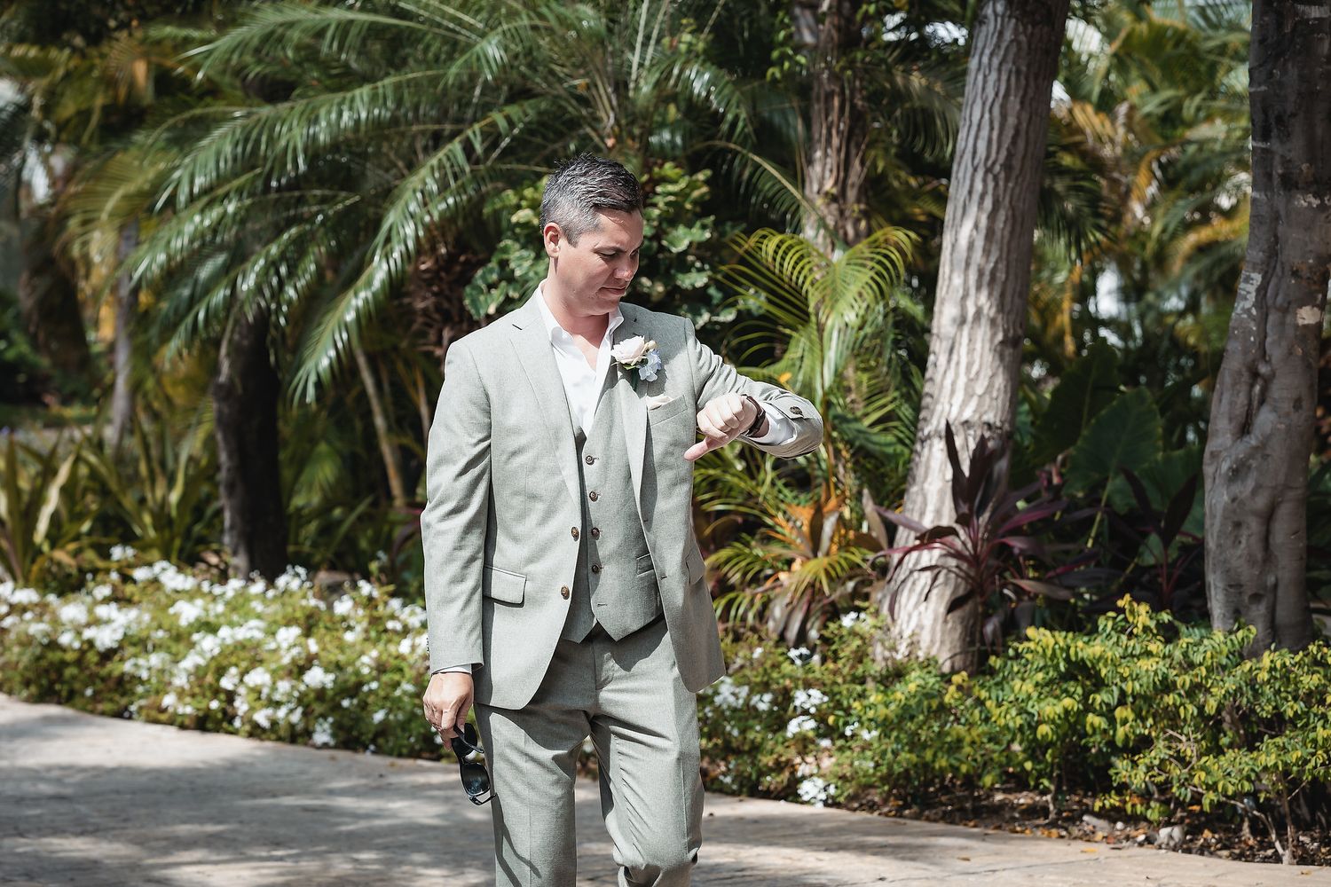Groom in light sage suit checking watch outdoors among tropical palms and white flowers at wedding venue.