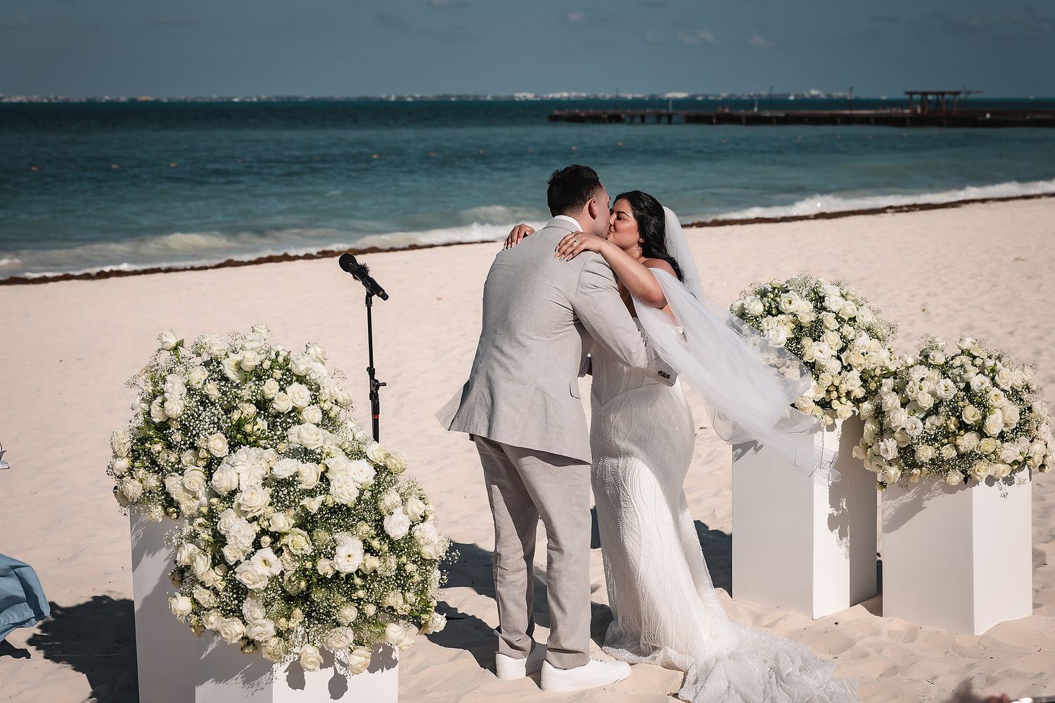 Couple sharing first kiss at romantic beach wedding ceremony with white floral arrangements and ocean backdrop.