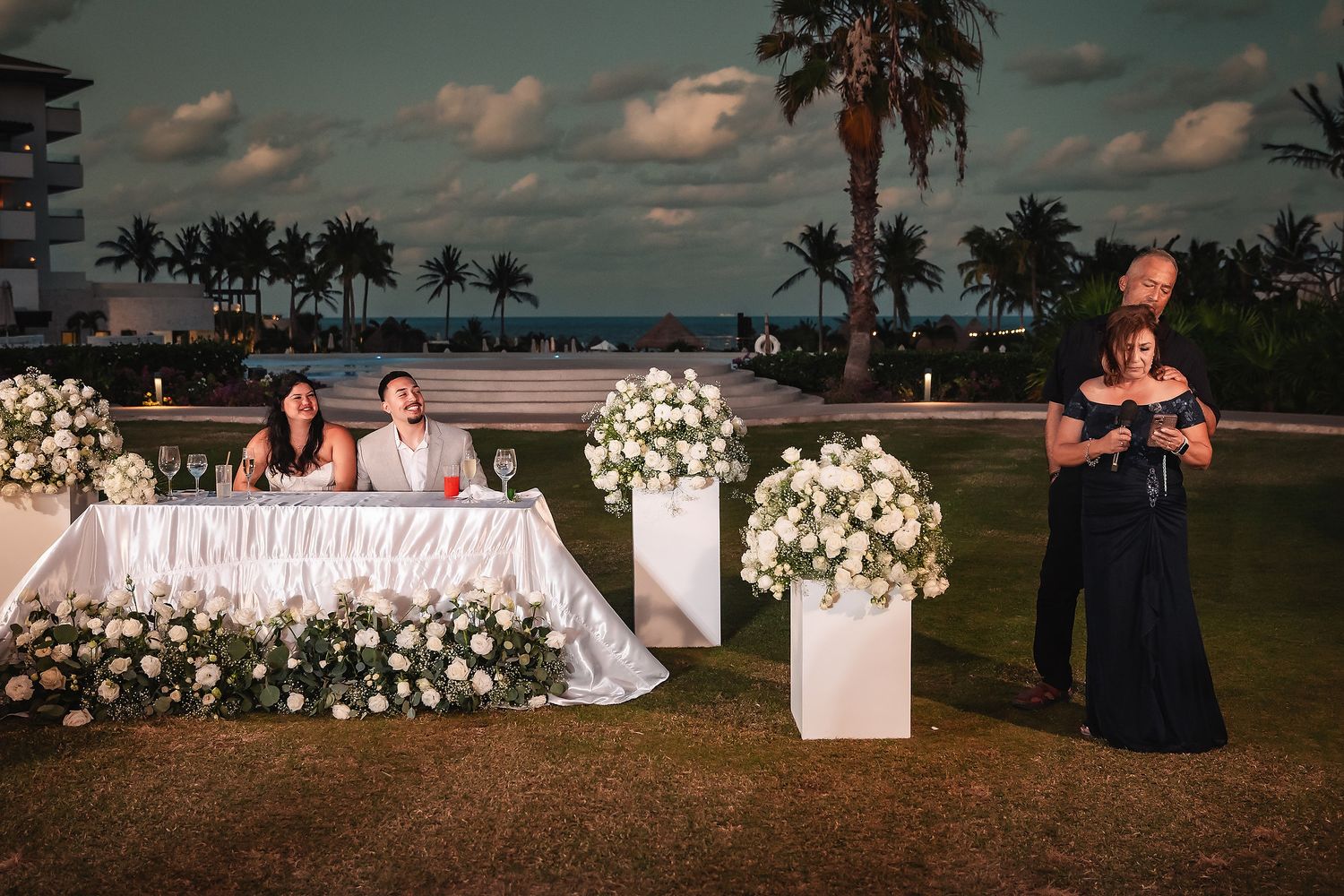 Elegant outdoor wedding reception at dusk with white floral arrangements, bridal party at decorated table, and palm trees near ocean.