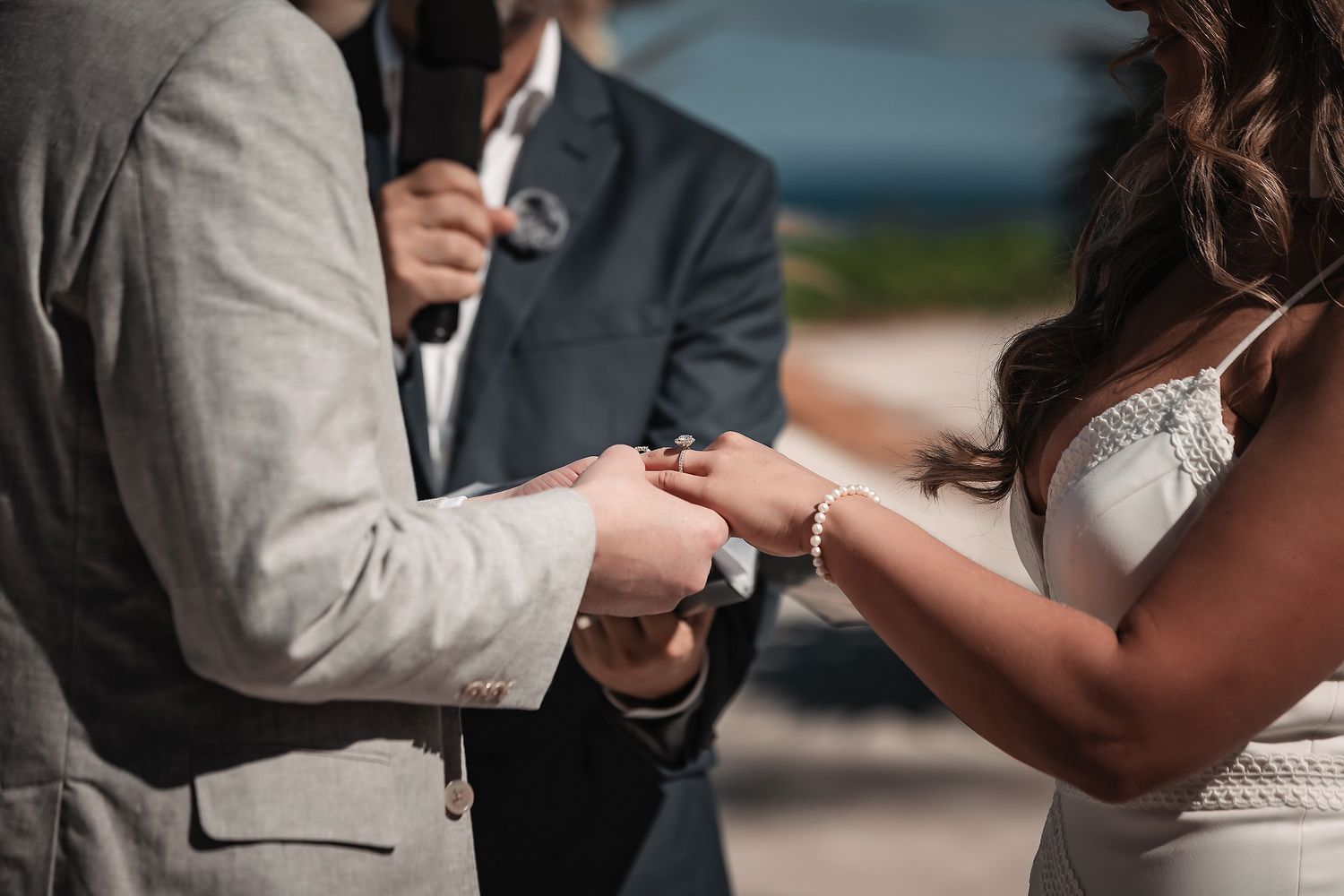 Groom placing wedding ring on bride's finger during outdoor ceremony, officiant in background.