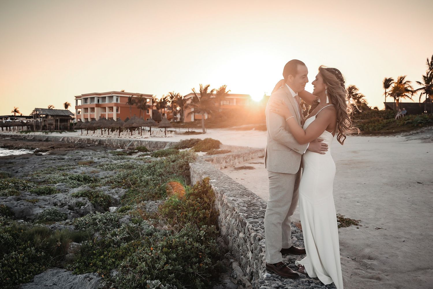Couple embraces on rocky beach at sunset during wedding photoshoot with tropical resort in background.