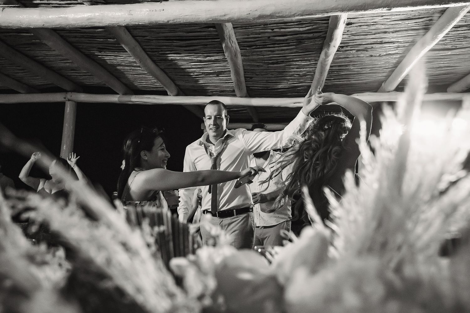 Black and white photo of guests dancing joyfully under a rustic wooden ceiling at an outdoor celebration.