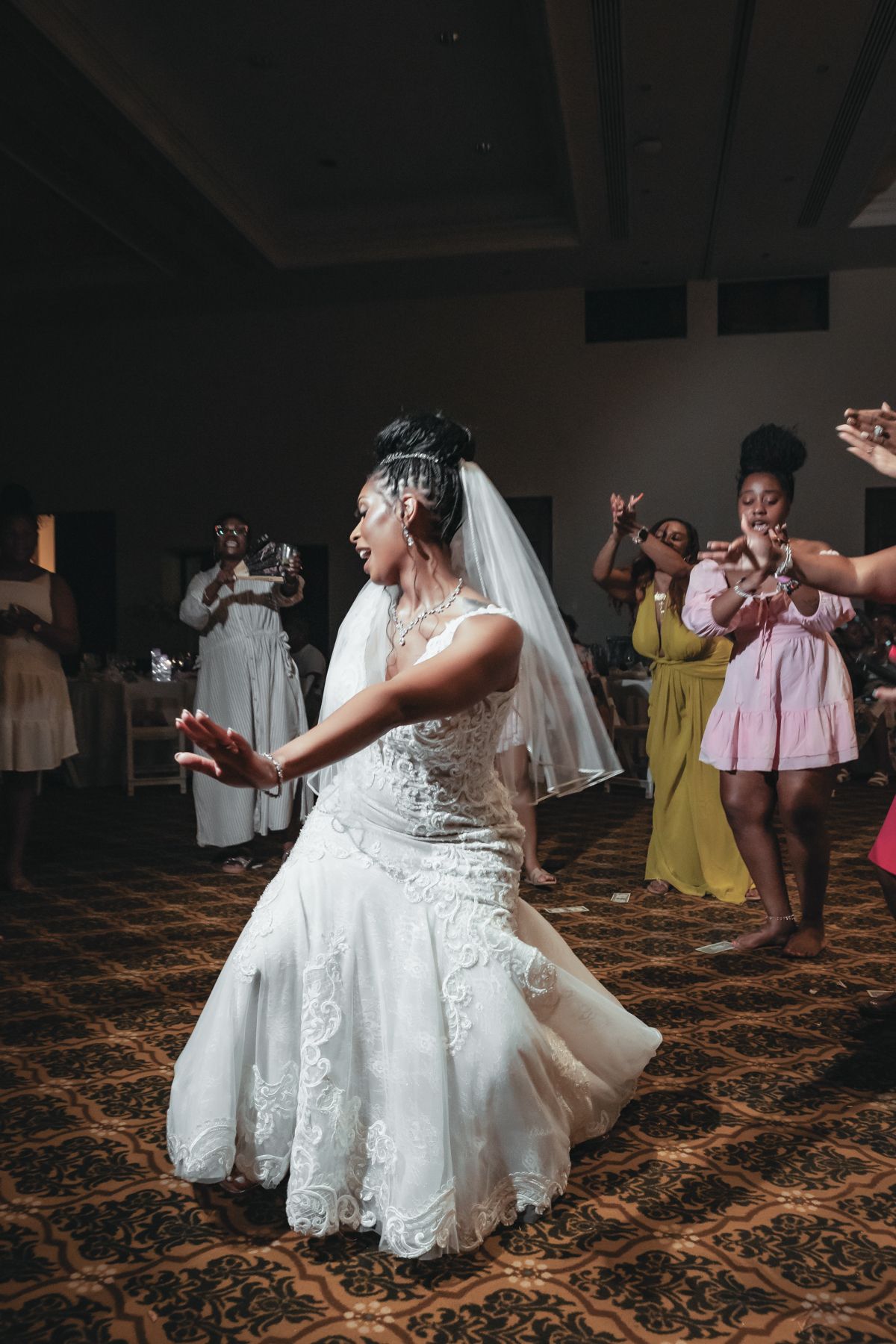 Bride in lace wedding dress and veil dancing joyfully at wedding reception with guests celebrating in background.