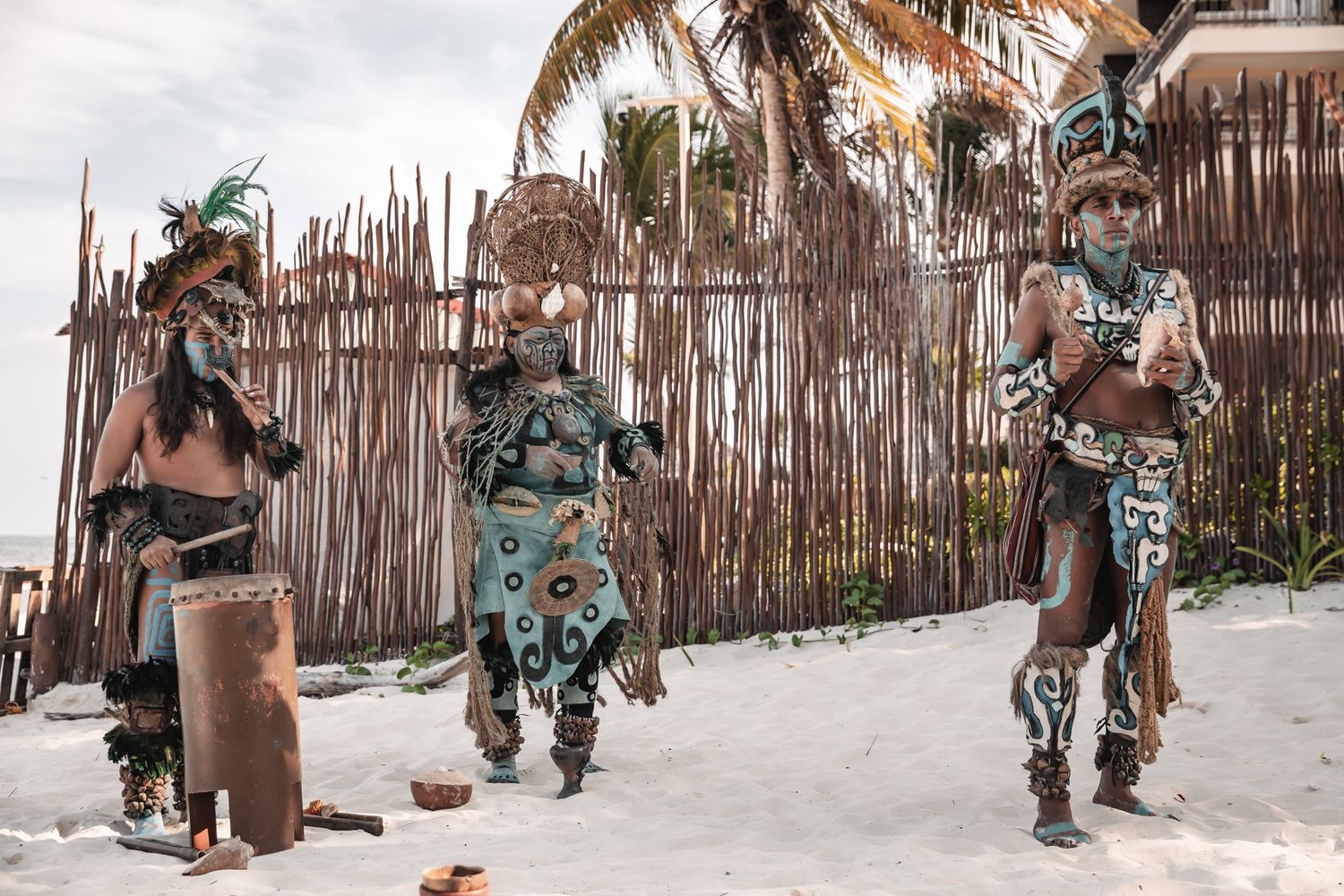 Three performers in traditional Mayan warrior costumes dance on a sandy beach near a bamboo fence.