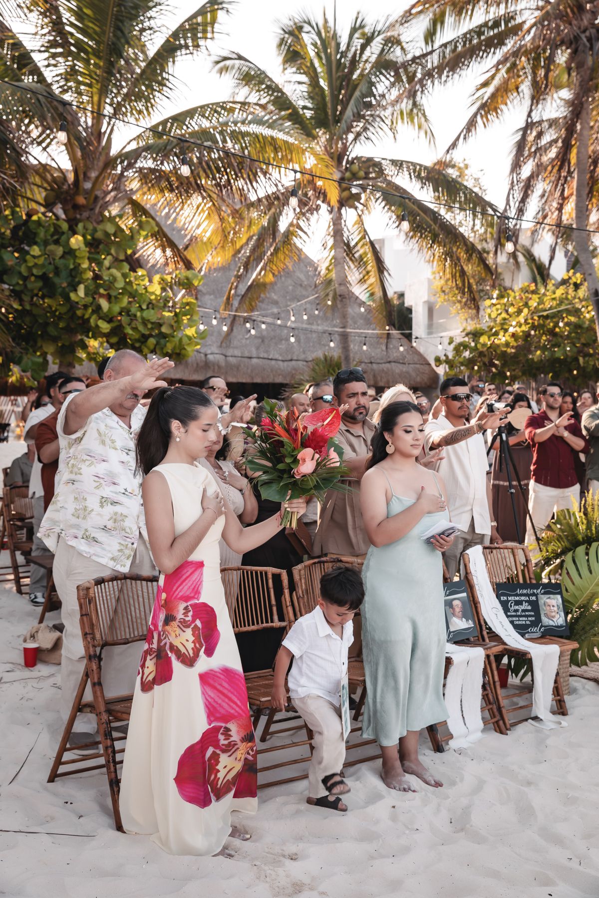 Wedding guests applauding on sandy beach at sunset, surrounded by palm trees and bamboo chairs in tropical setting.