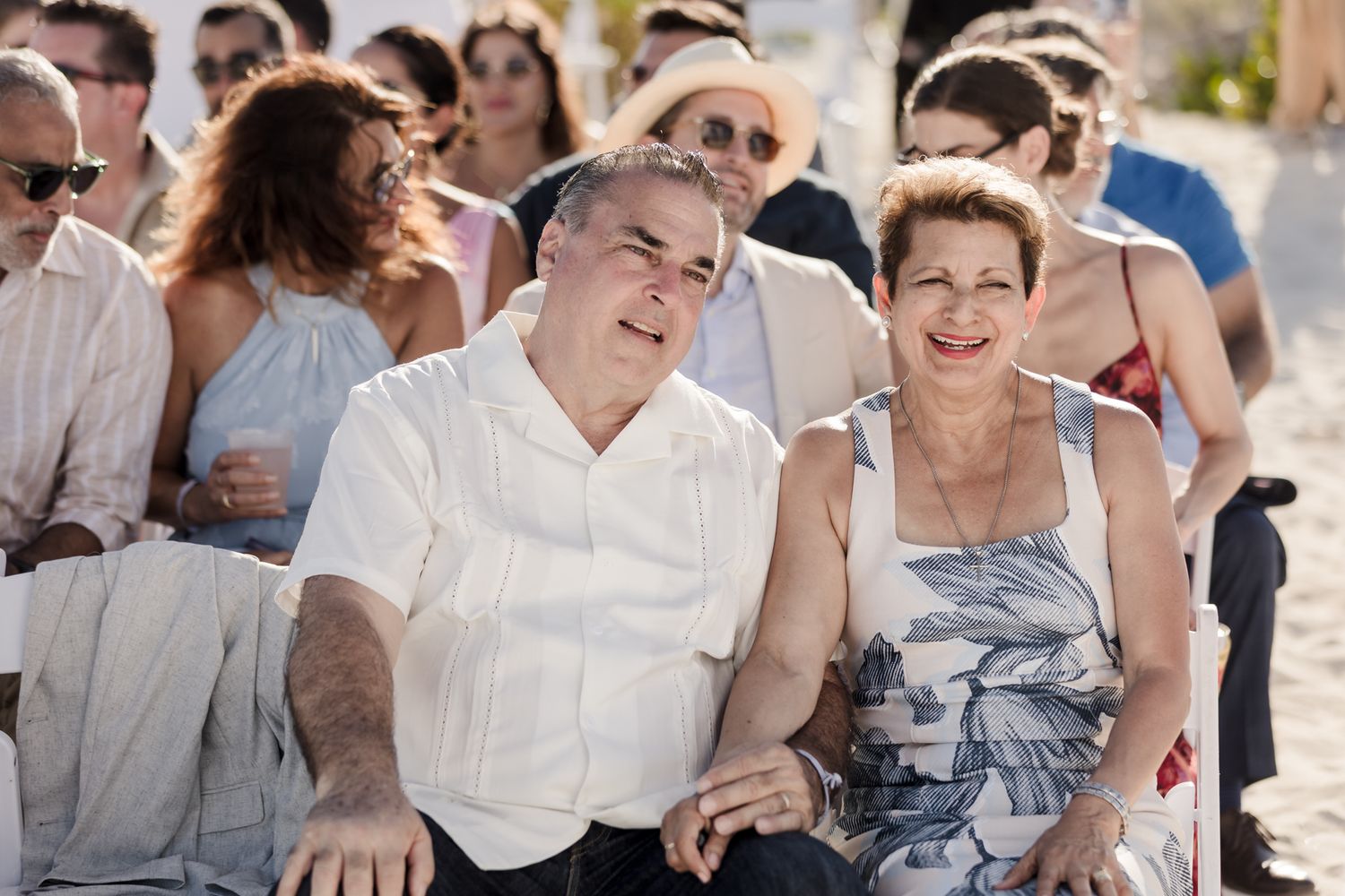 Smiling couple sitting together at an outdoor beach wedding ceremony with guests in the background.