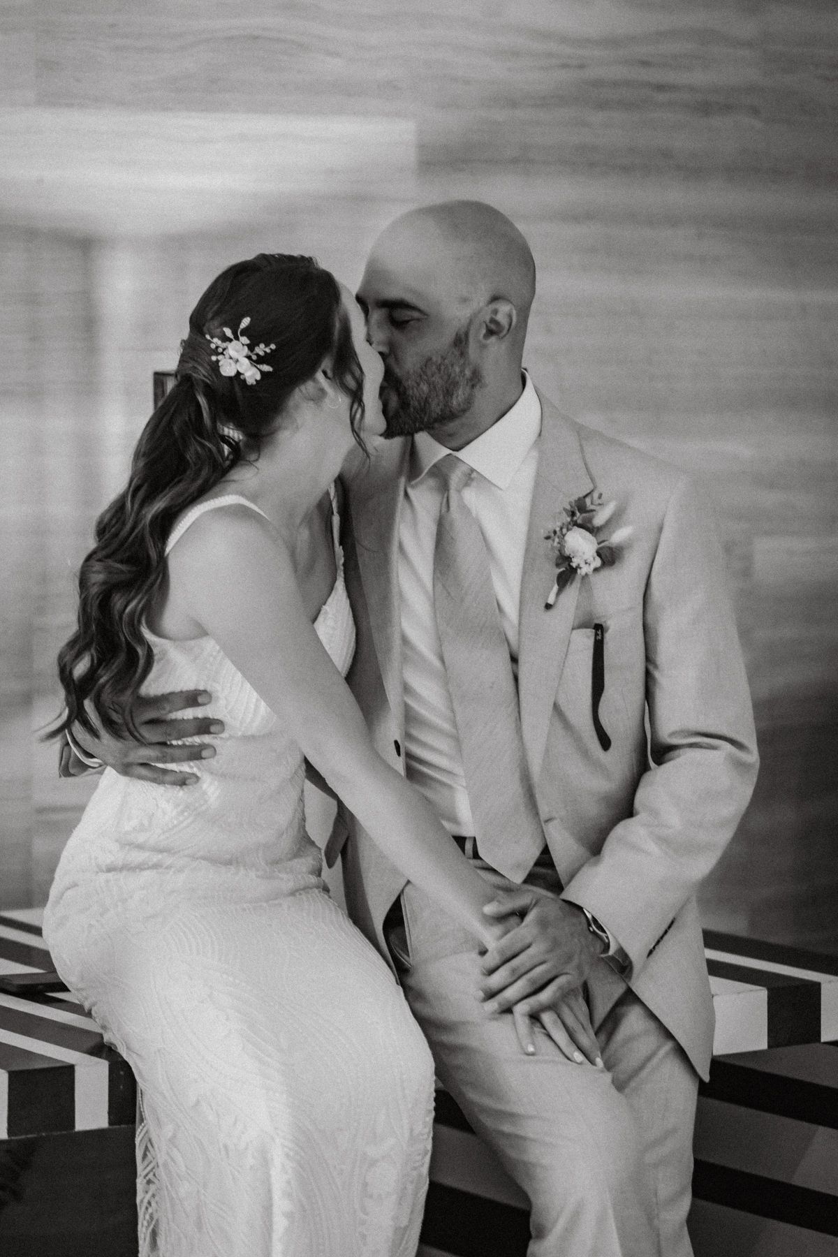 Black and white photo of a bride and groom sharing a tender kiss while seated, groom in suit with boutonniere.