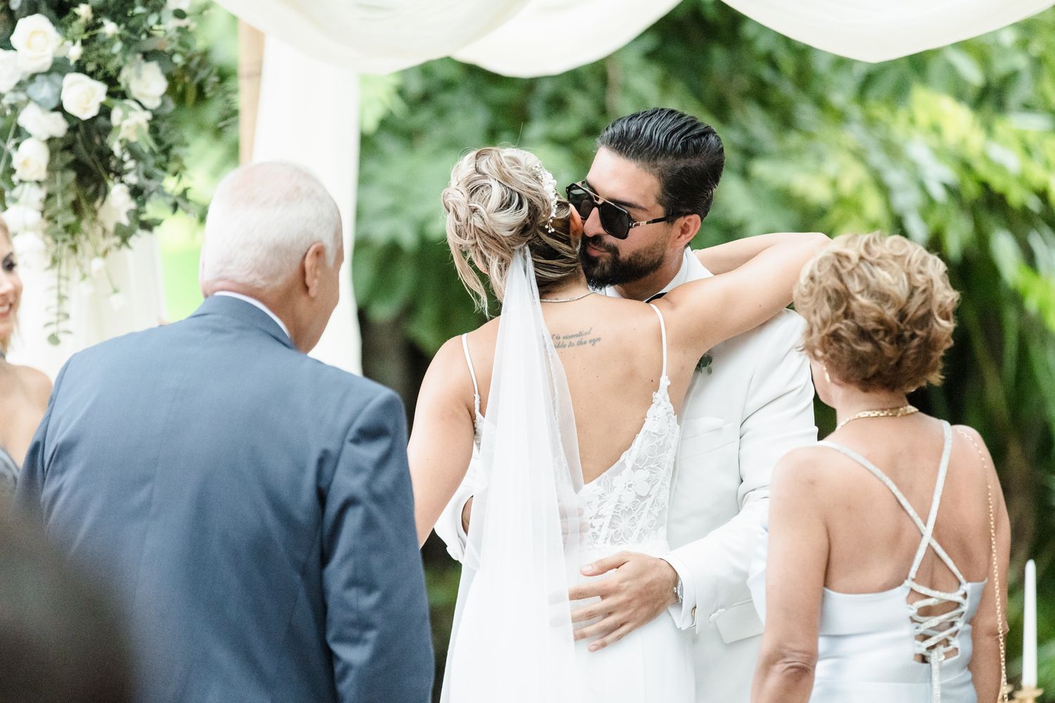 Bride and groom share a tender kiss at outdoor wedding ceremony surrounded by family and floral arch.