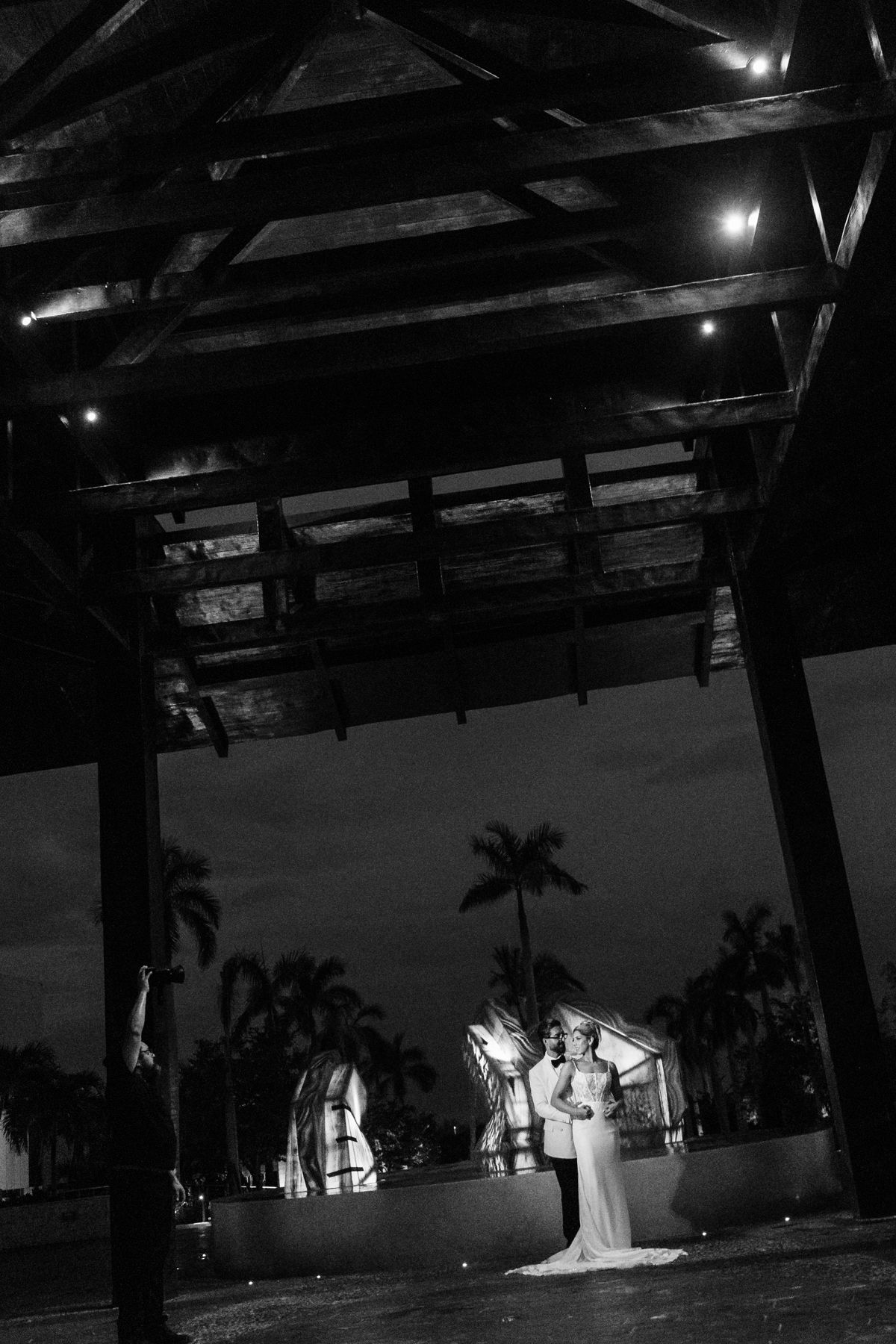 Black and white night wedding photo of bride and groom under wooden pavilion with tropical palm trees.