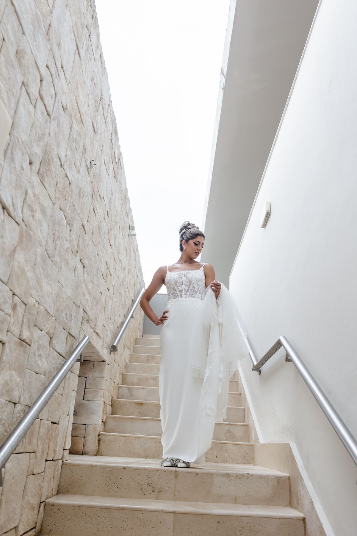 Bride in white lace gown descending marble stairs, holding veil, wearing elegant updo hairstyle.