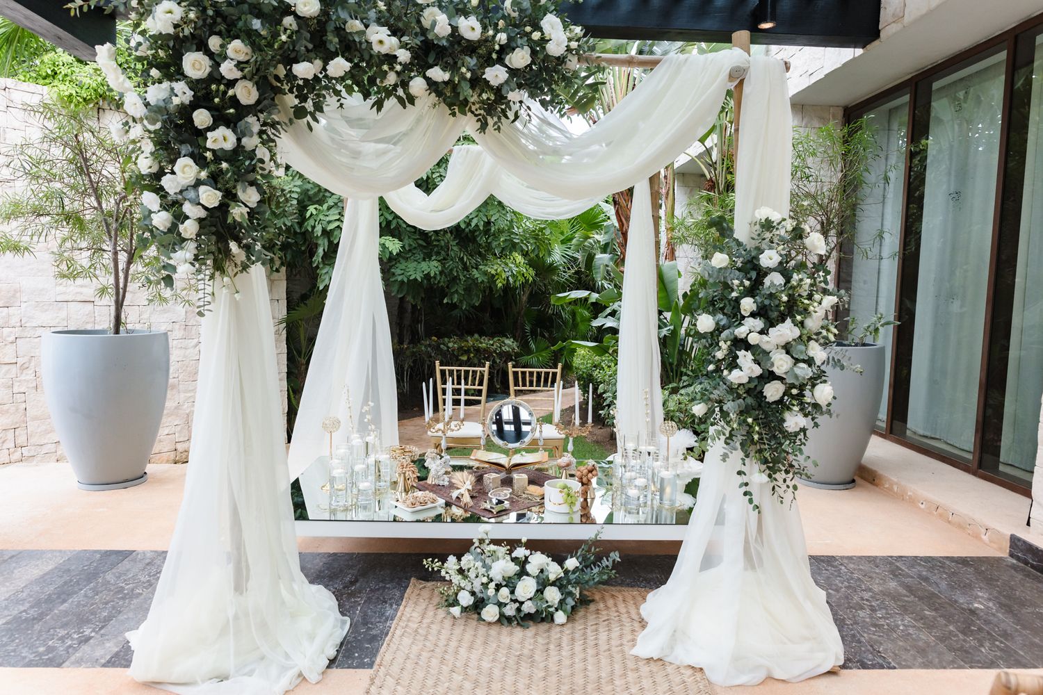 Elegant outdoor wedding ceremony setup with white floral arch, draped fabric, and decorated table surrounded by lush greenery.