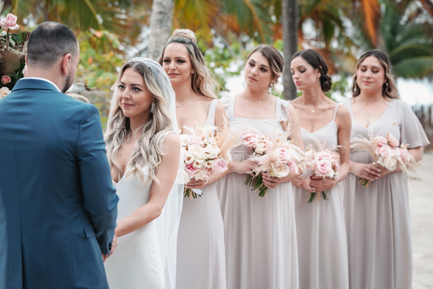 Bride in white gown with veil stands at altar with bridesmaids holding pink bouquets on beach.