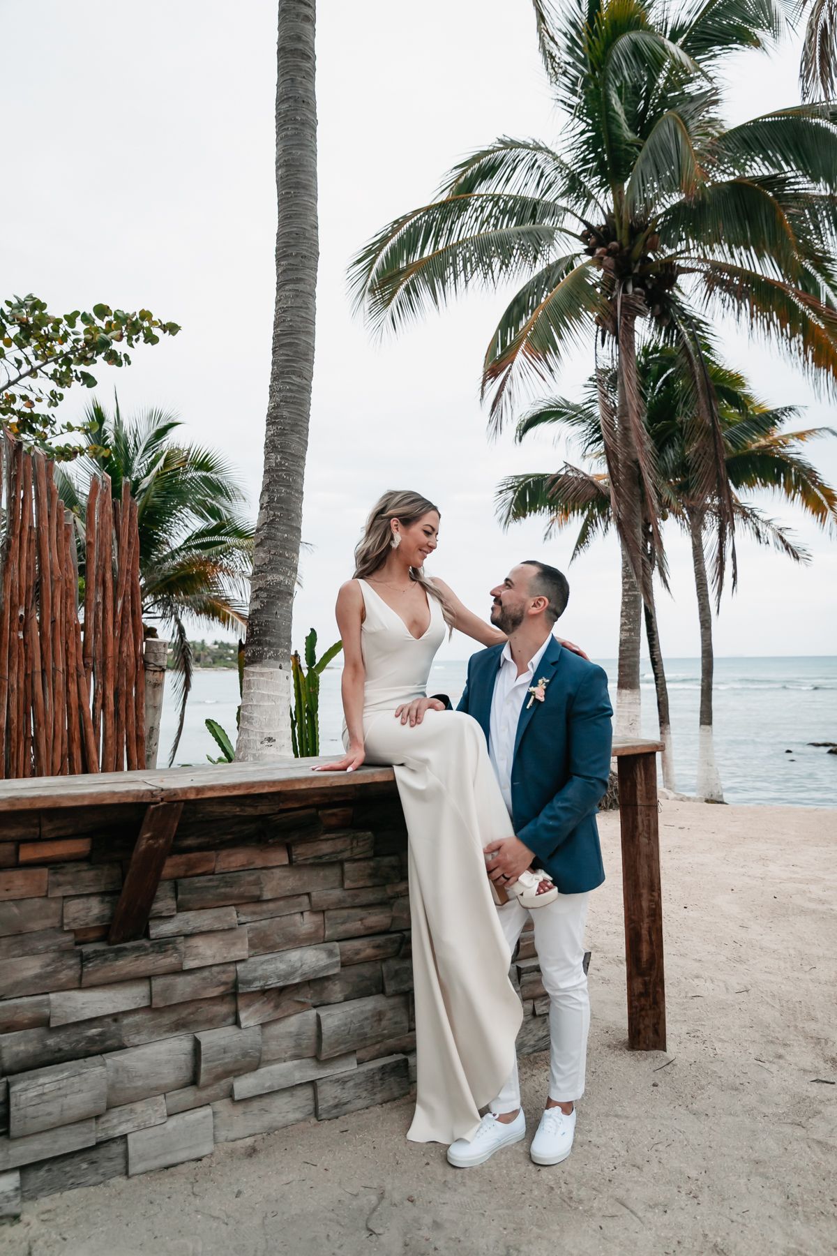 Bride in white gown sits on stone ledge as groom in blue suit gazes at her among tropical palms.