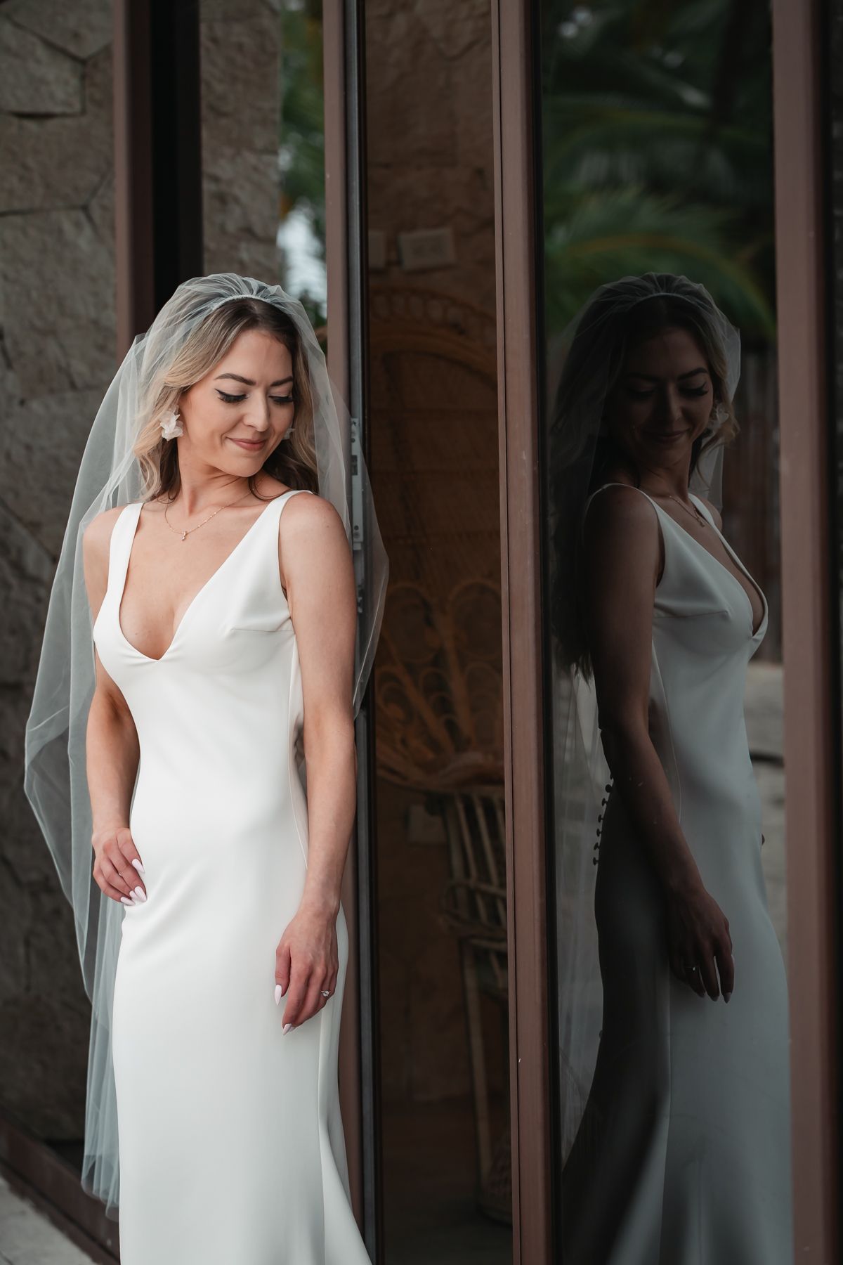 Elegant bride in a sleek white satin gown and veil poses beside a mirror reflection at her wedding.