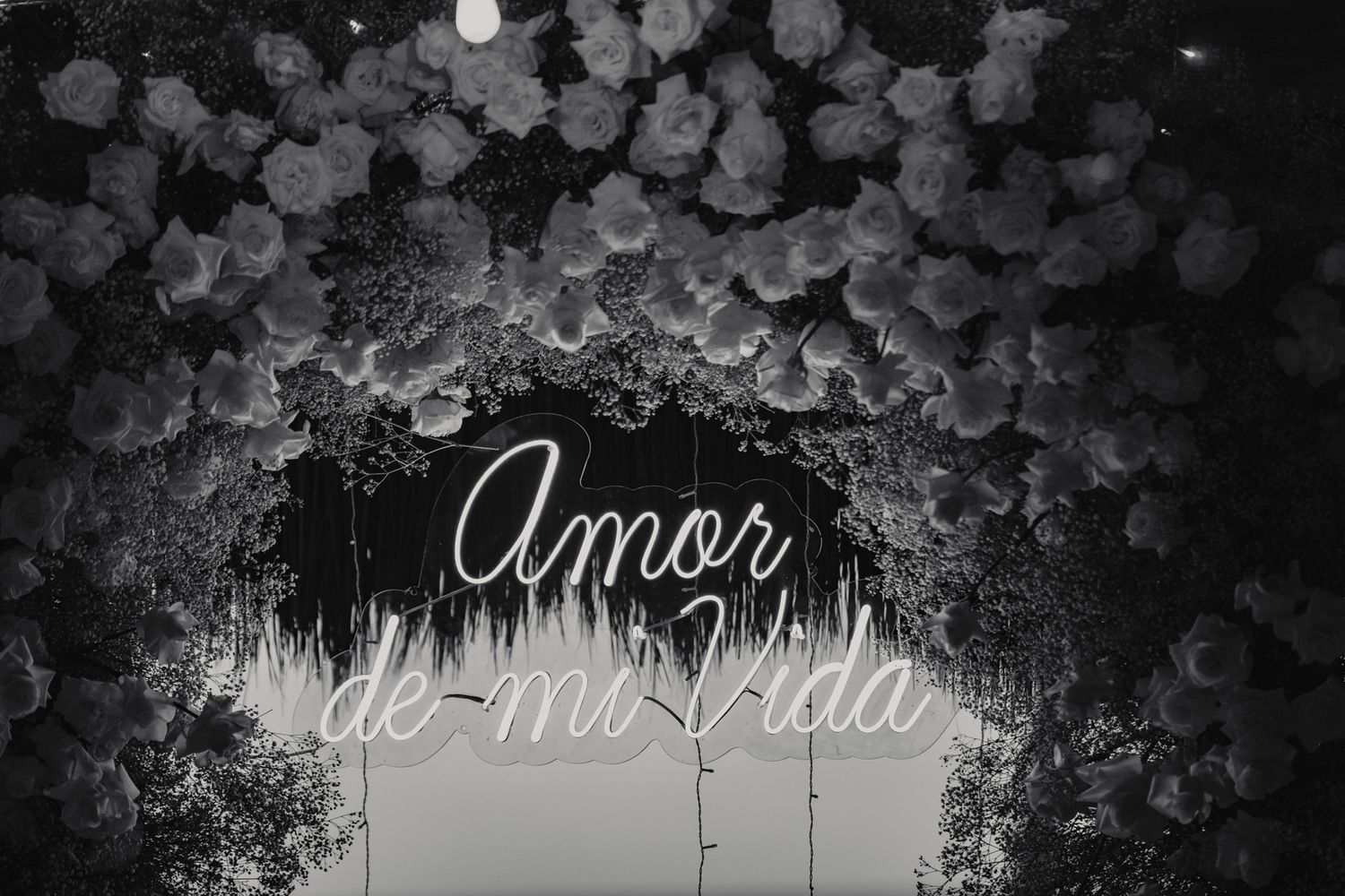 Black and white photo of a floral arch with neon sign reading Amor de mi Vida at a wedding.