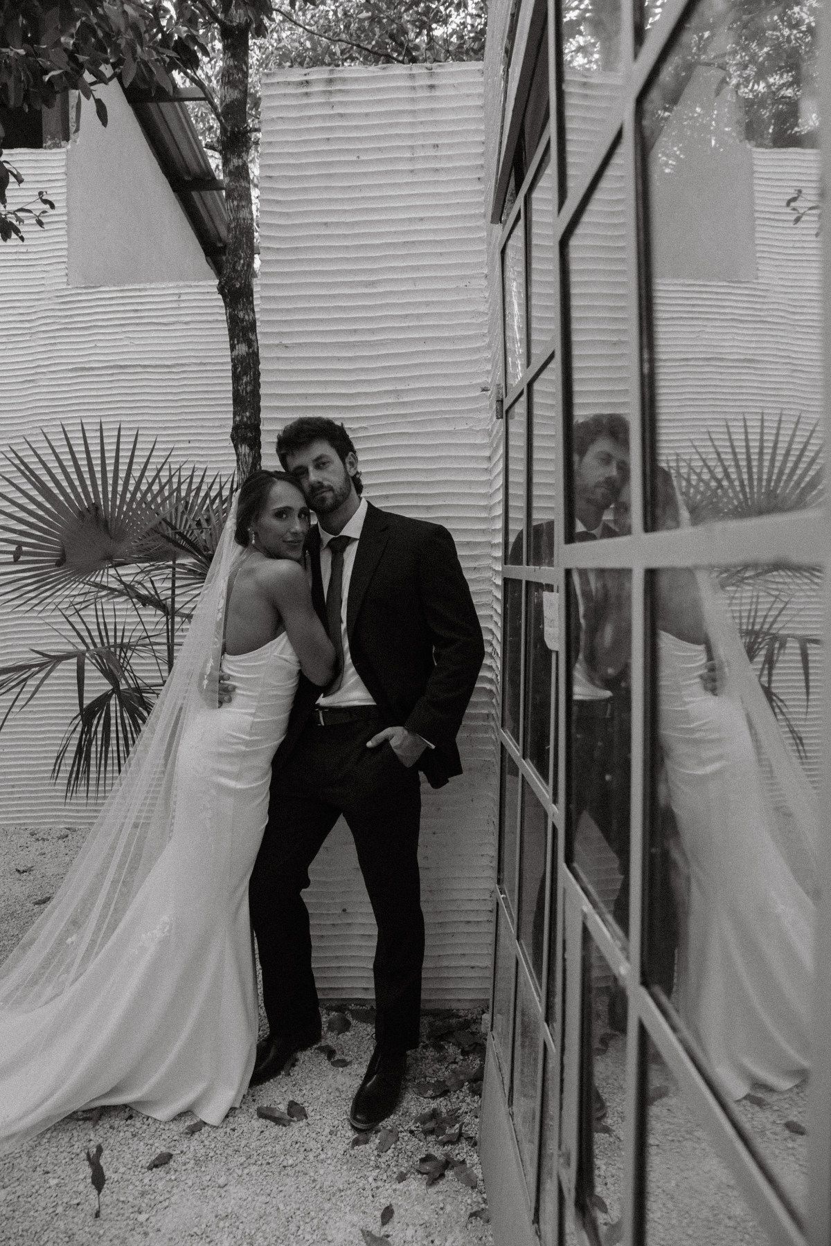 Black and white photo of a bride and groom posing elegantly by a glass door with tropical plants in background.