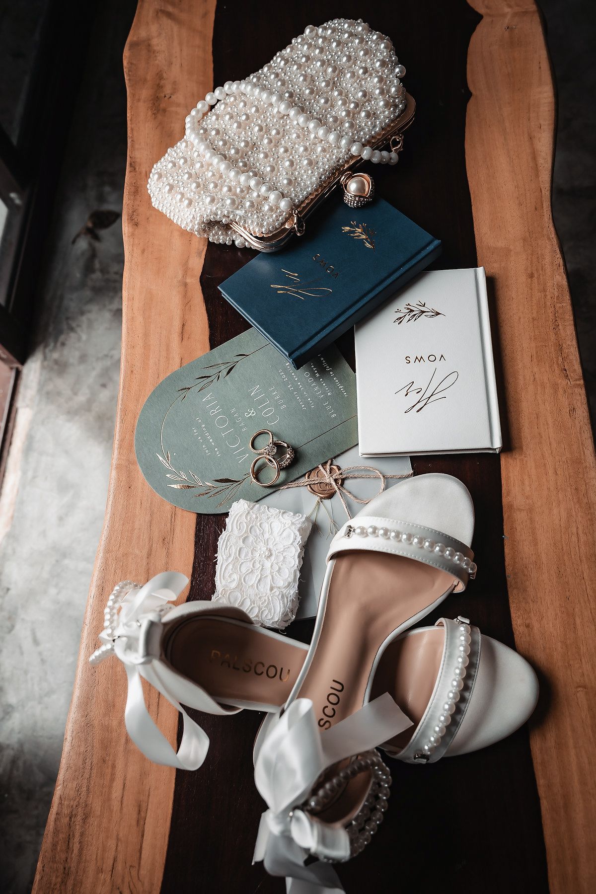 Elegant bridal flat lay featuring pearl heels, pearl clutch, wedding rings, vows booklet, and sage green invitation on wood.