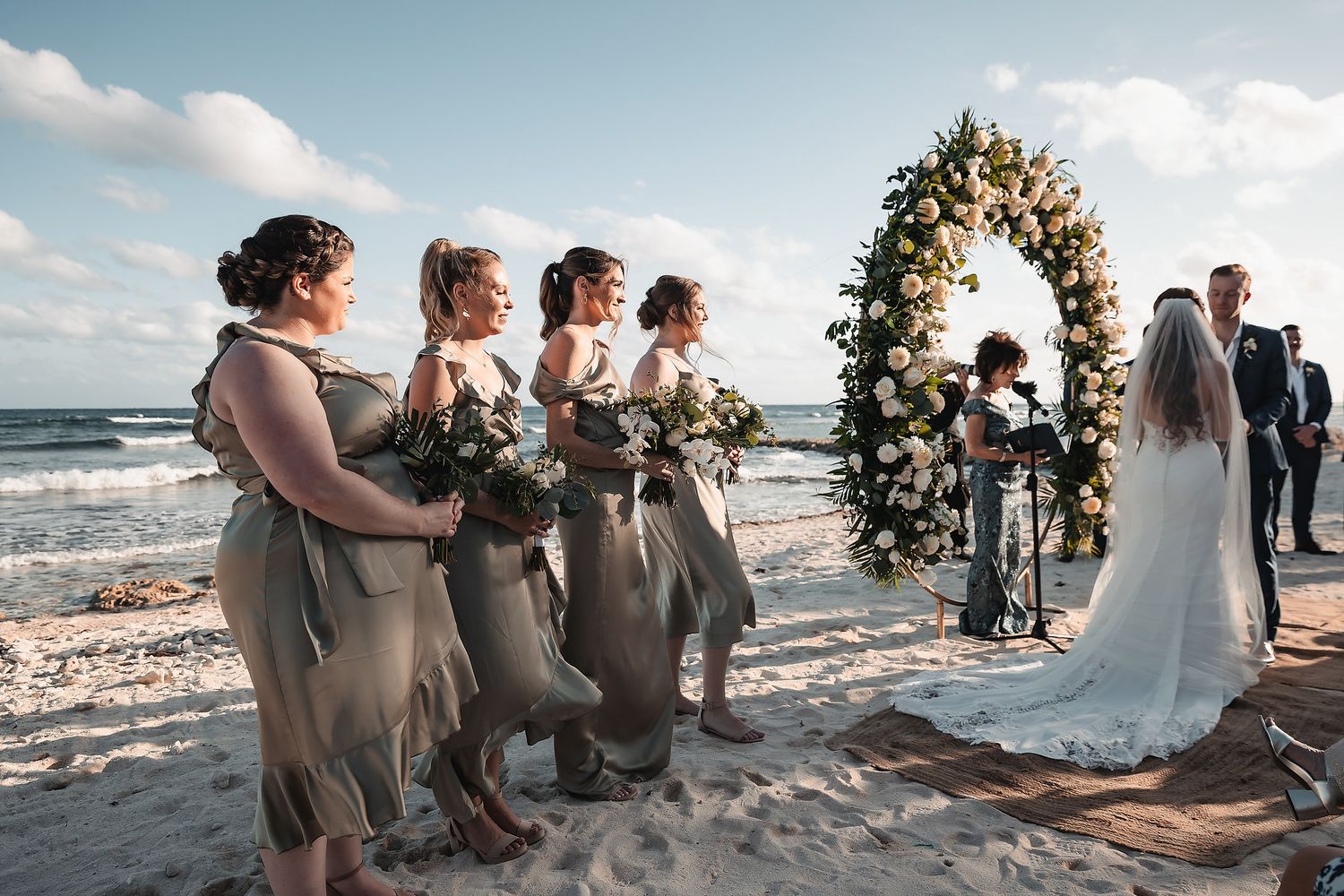 Bridesmaids in olive dresses hold bouquets at a beach wedding ceremony with a floral arch and ocean backdrop.