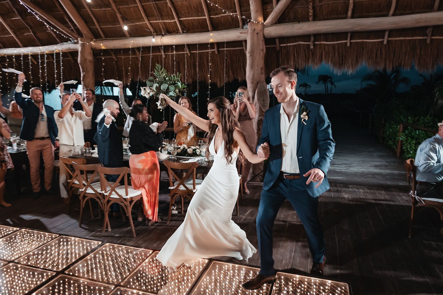 Bride and groom share a joyful first dance at an outdoor wedding reception with string lights and guests celebrating.
