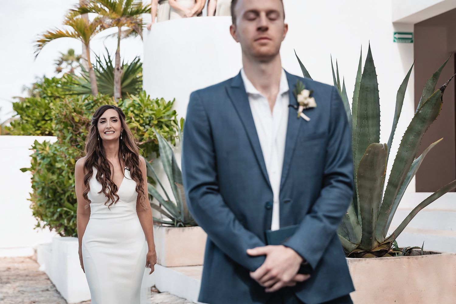 Bride in white gown smiles at groom in blue suit during outdoor first look at tropical wedding venue.
