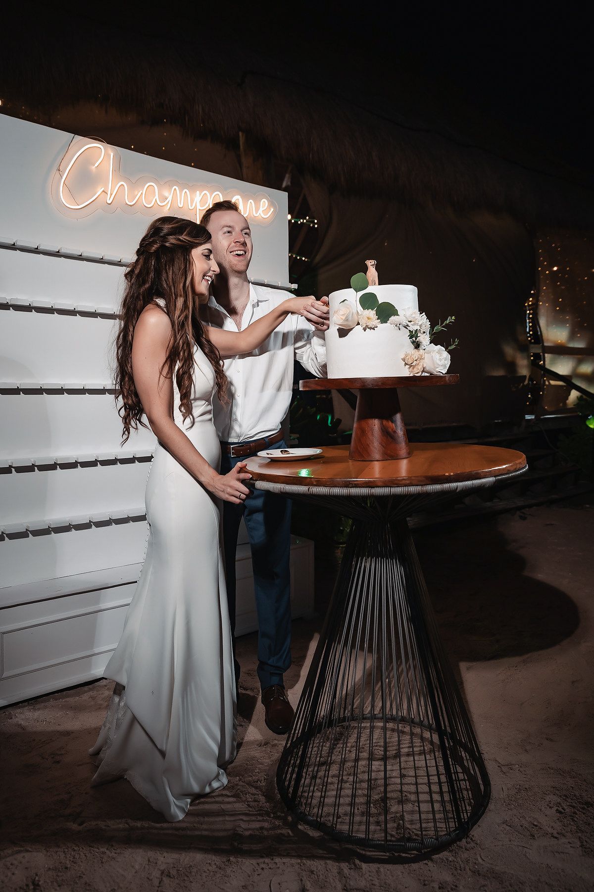 Bride and groom cutting their white floral wedding cake at a reception with a glowing Champagne neon sign backdrop.