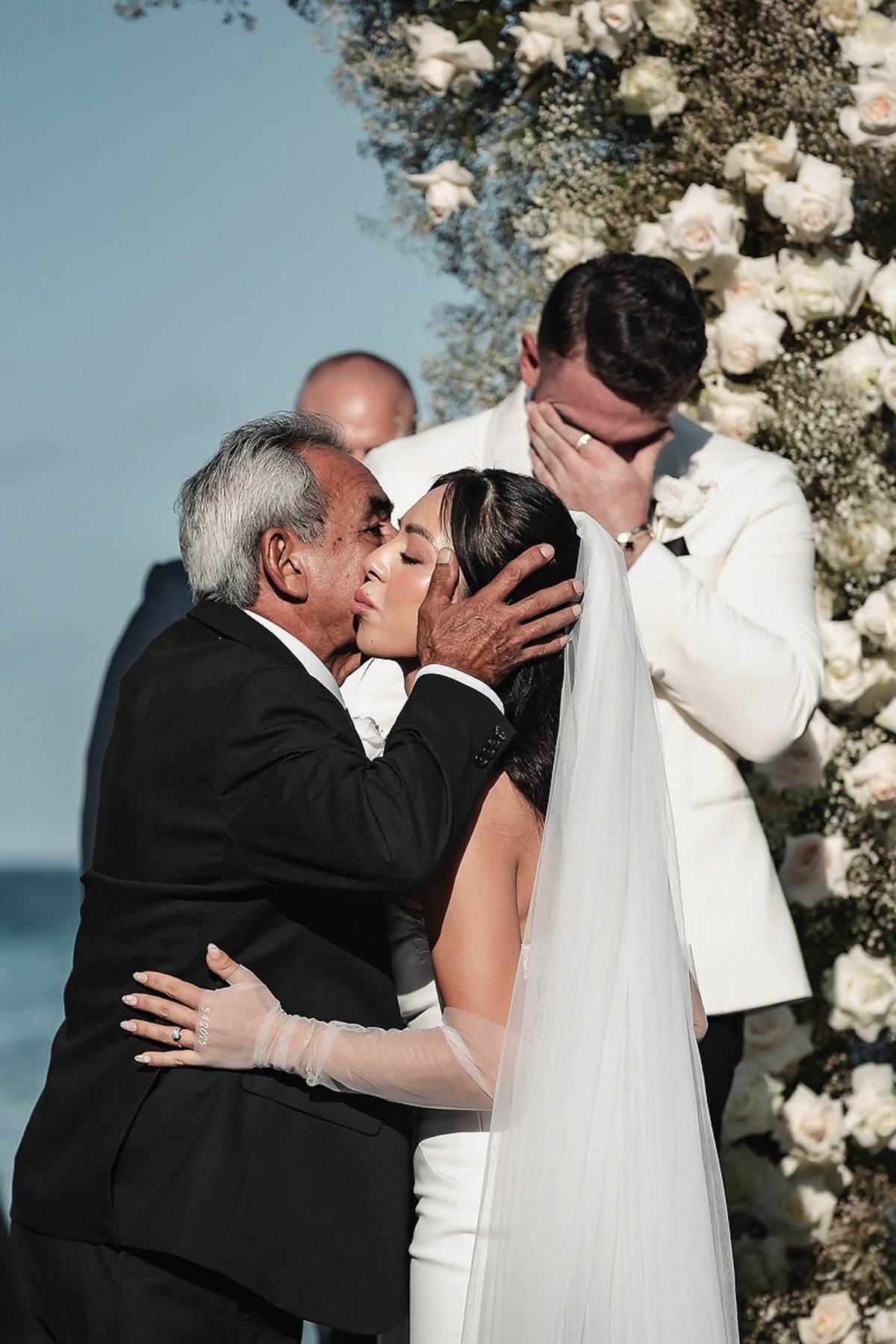 Father of the bride kisses bride as emotional groom covers face during outdoor wedding ceremony with floral arch.