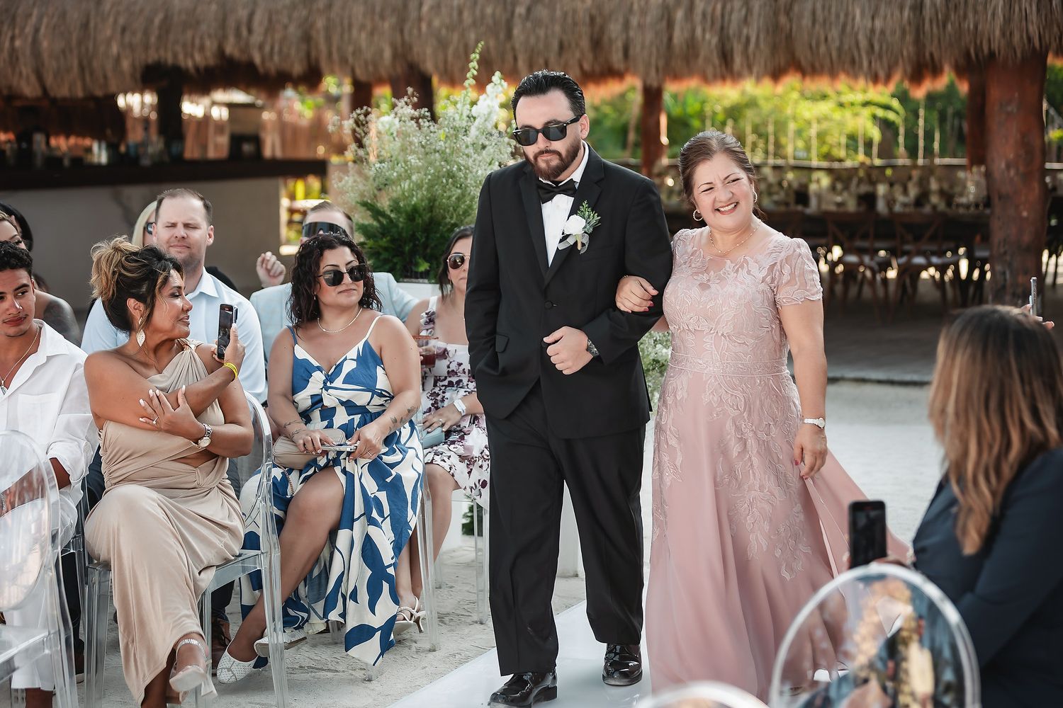 Groom in tuxedo walks down wedding aisle with smiling woman in pink gown, guests seated alongside.