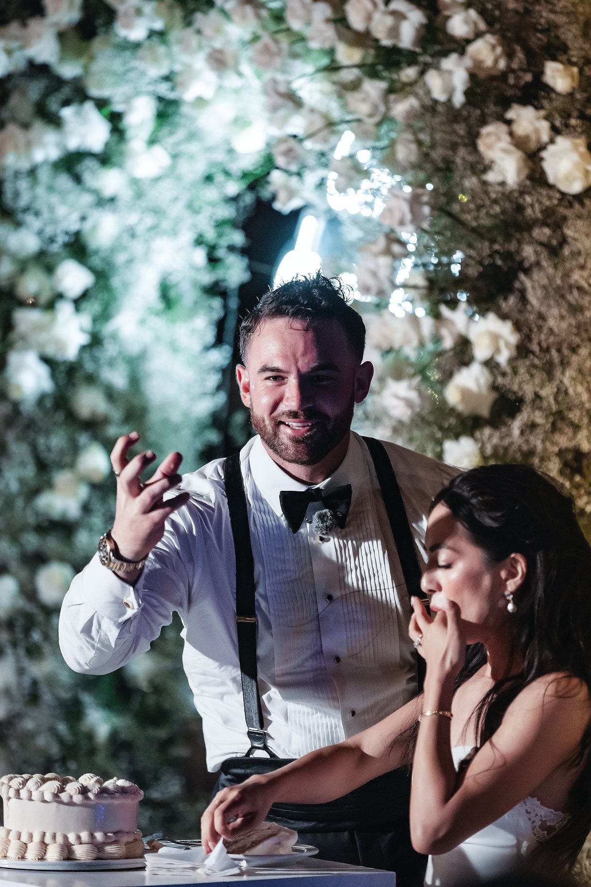 Groom in suspenders and bow tie laughs while bride tastes wedding cake at floral-decorated reception.
