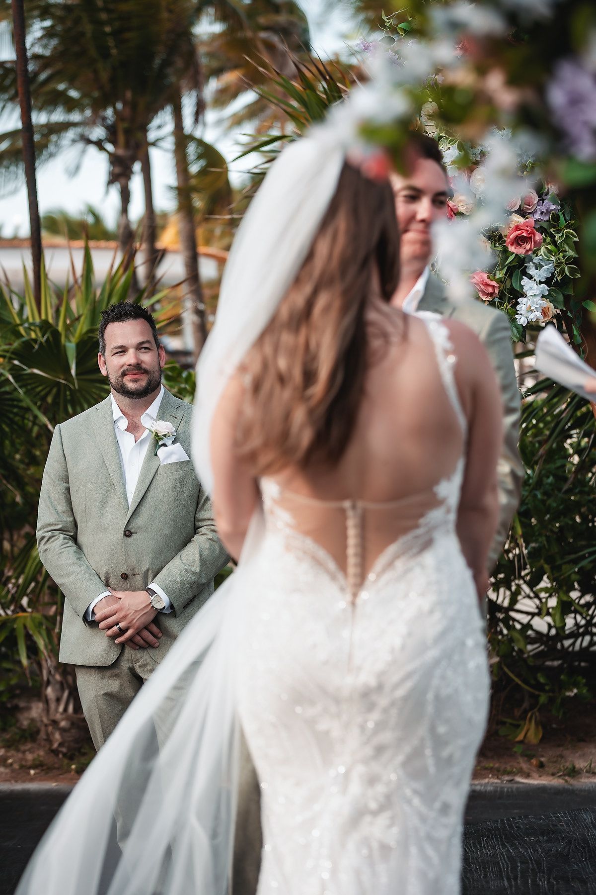 Groom in sage suit watches bride in lace backless gown walk down aisle at outdoor tropical wedding ceremony.