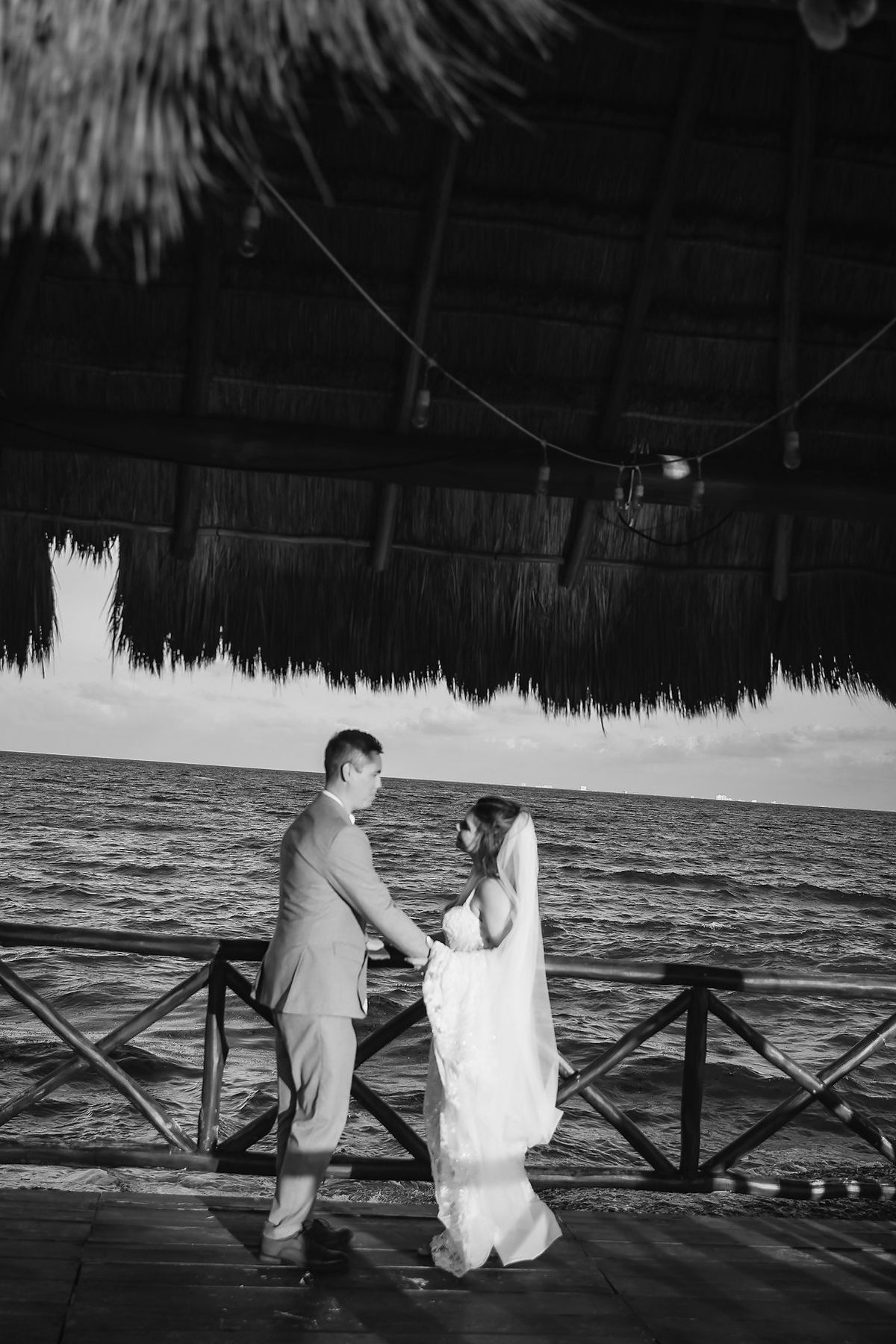Black and white photo of a bride and groom holding hands on a seaside deck under a thatched roof.