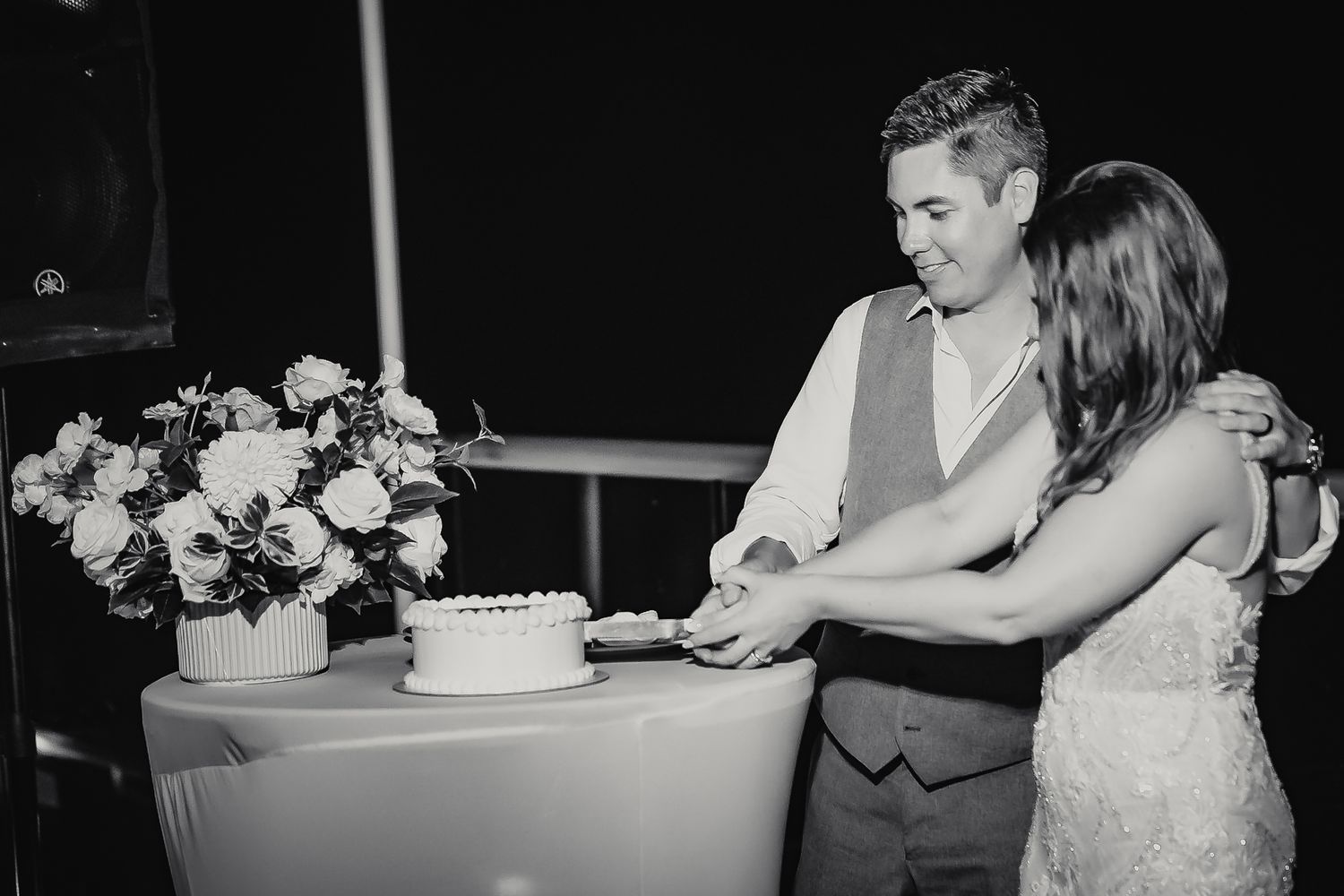 Couple cutting their wedding cake together in black and white photo with floral arrangement on table.