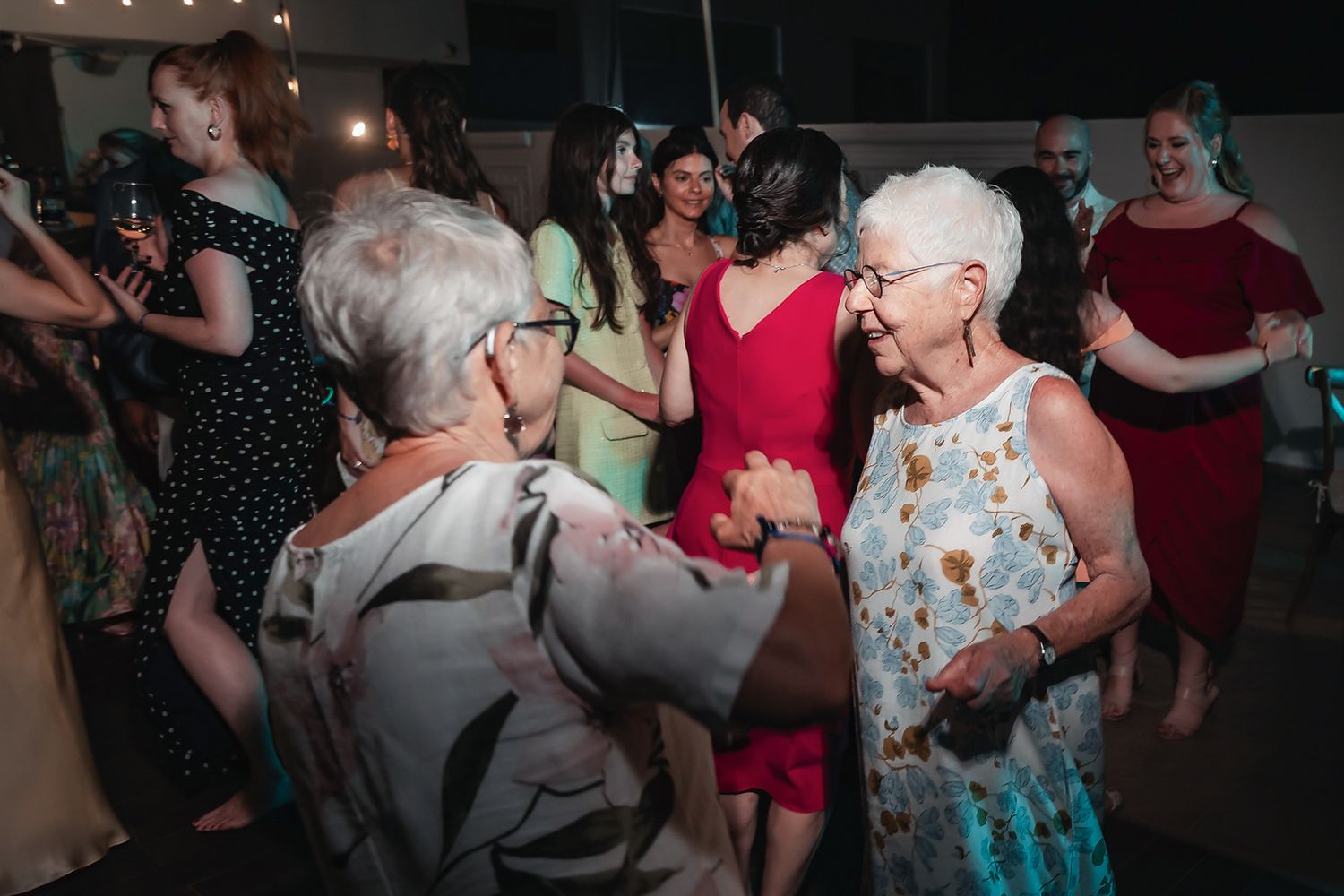 Elderly women dancing joyfully at a lively indoor party with guests in colorful dresses.
